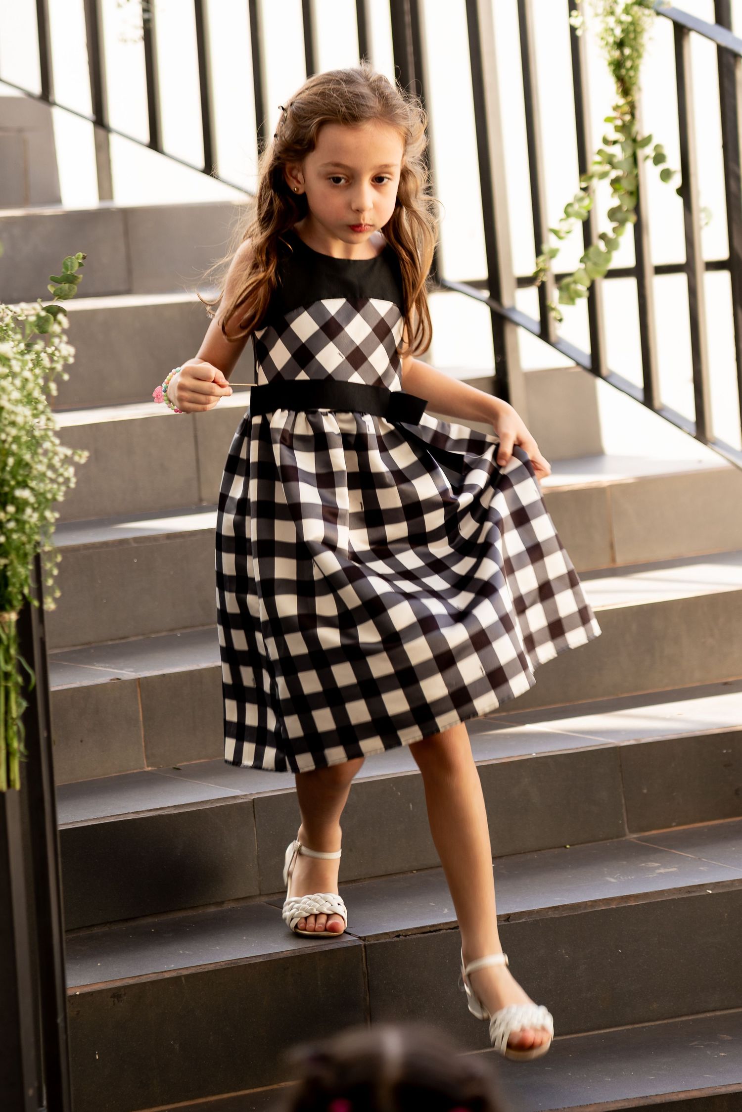 A young girl in a black and white gingham dress with a sash walks down stone steps wearing white sandals.