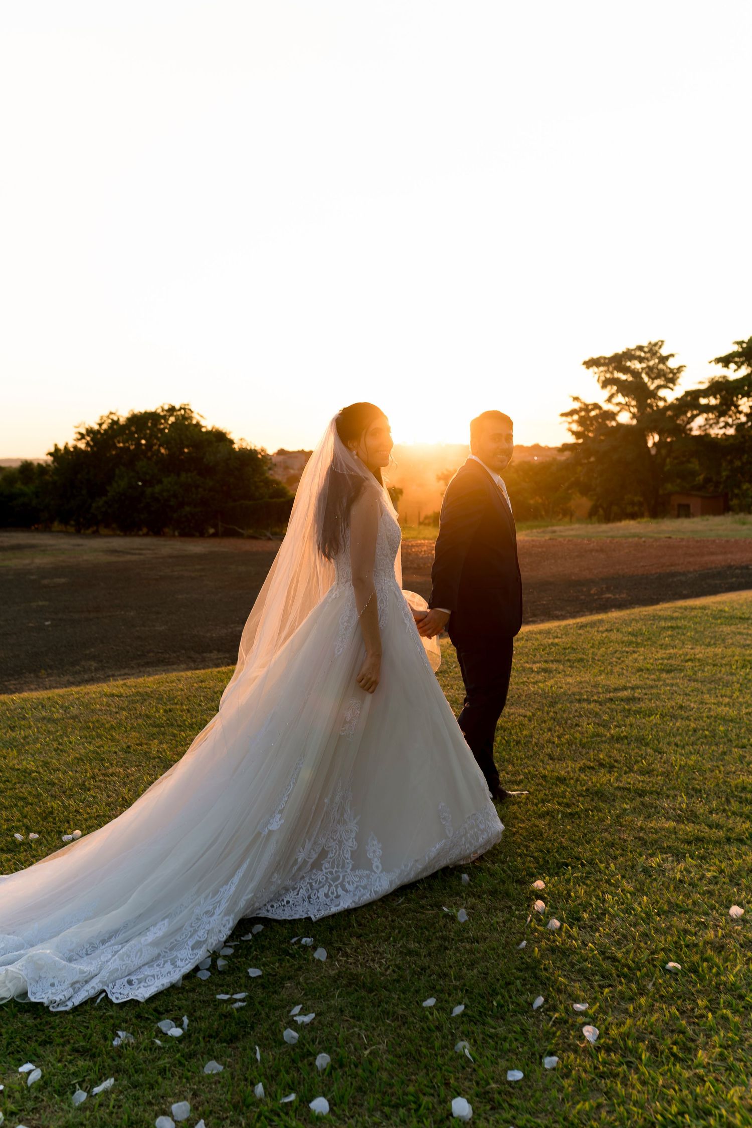 Bride and groom walk hand in hand on green lawn at golden sunset, her cathedral veil trailing behind.