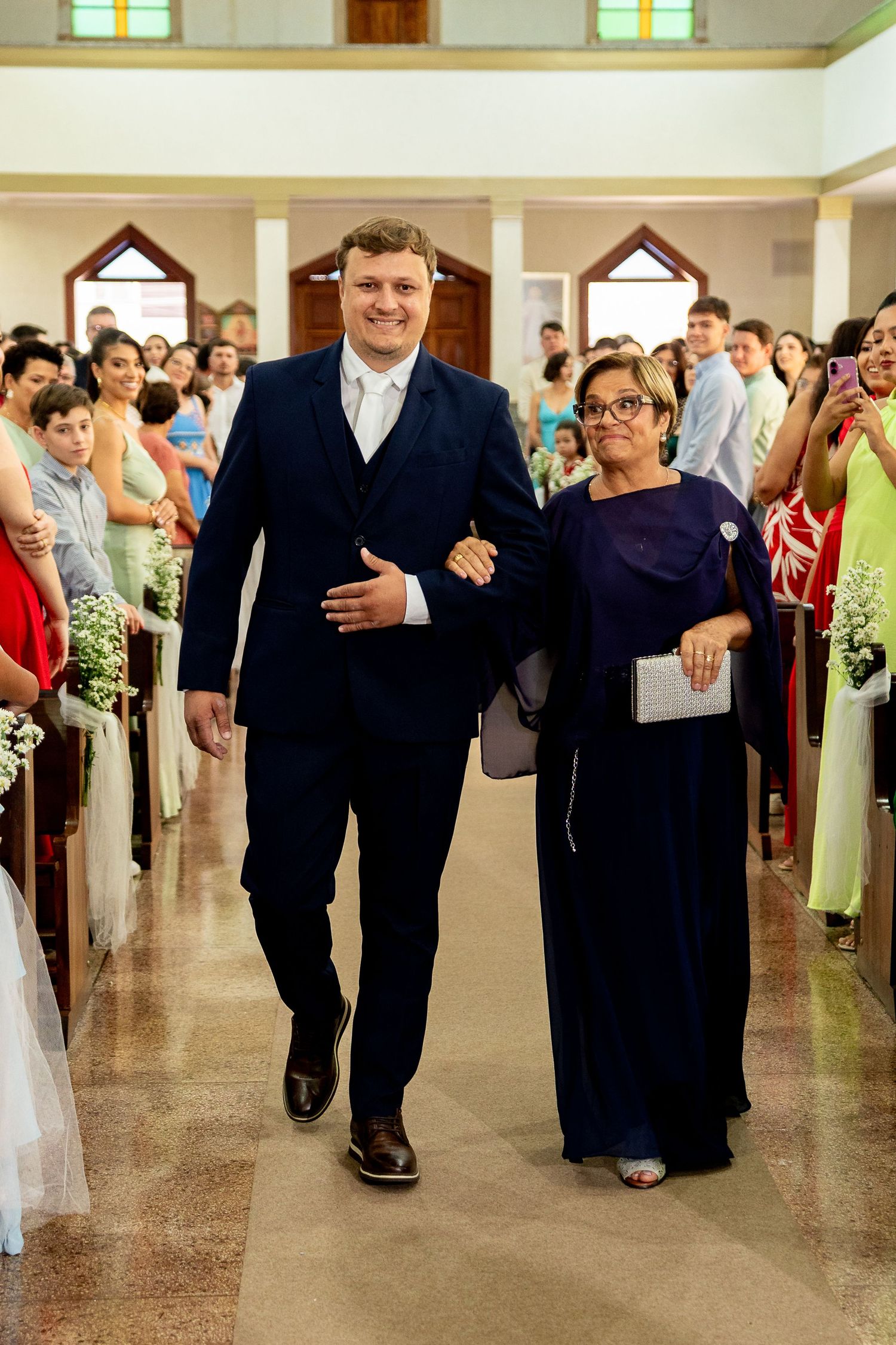 Groom in navy suit escorts woman in purple gown down church aisle at wedding ceremony.