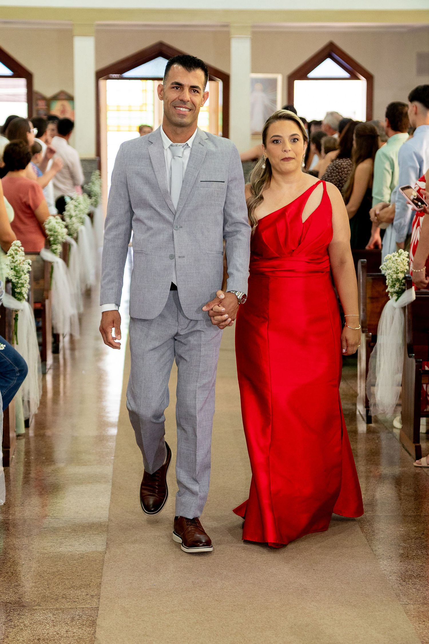 Couple walking down church aisle, man in gray suit, woman in elegant red gown, wedding ceremony guests seated.
