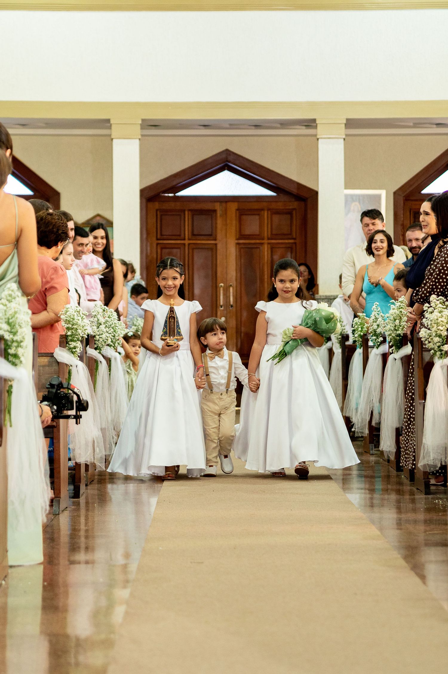 Flower girls in white dresses and ring bearer walk down church aisle at wedding ceremony.