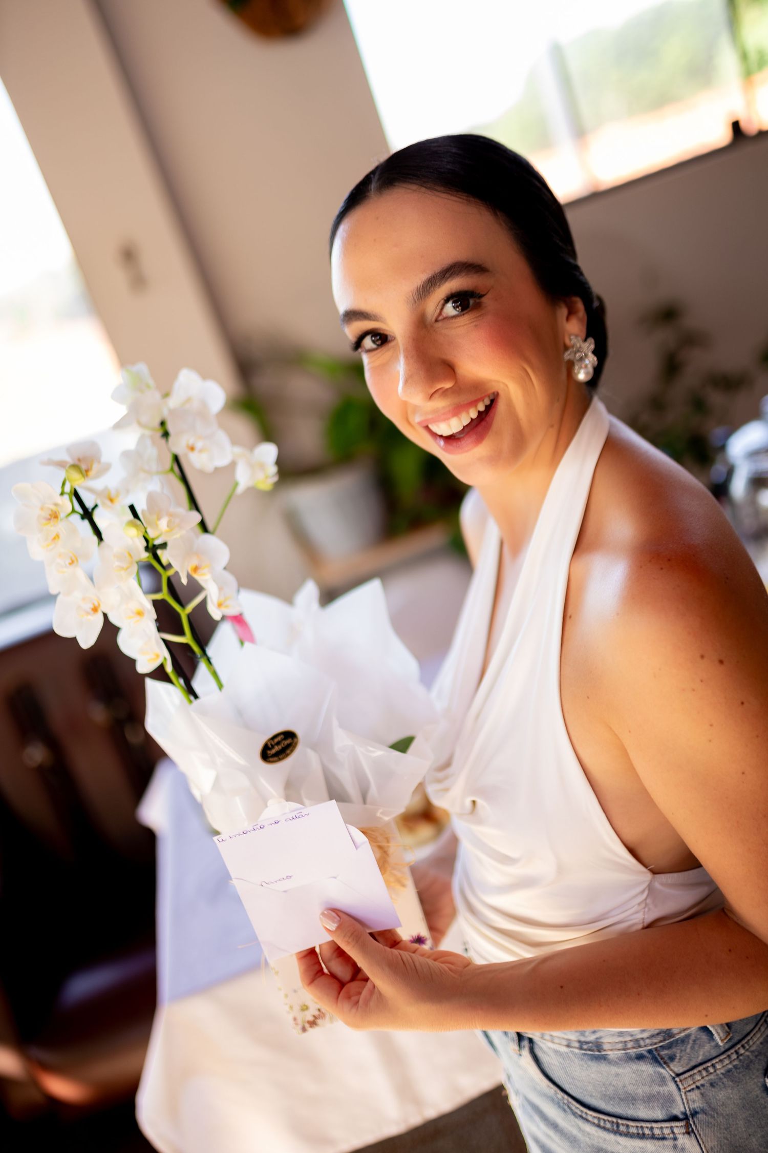 Smiling woman in white halter top holding white orchid flowers and a card indoors.