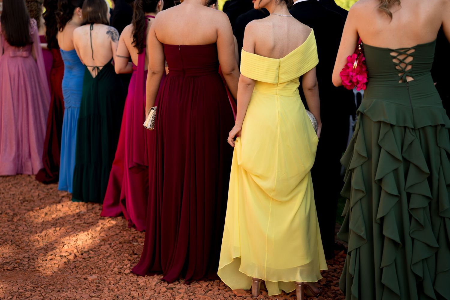 Women in colorful formal gowns including yellow, burgundy, and green stand in a row at a prom or formal event.