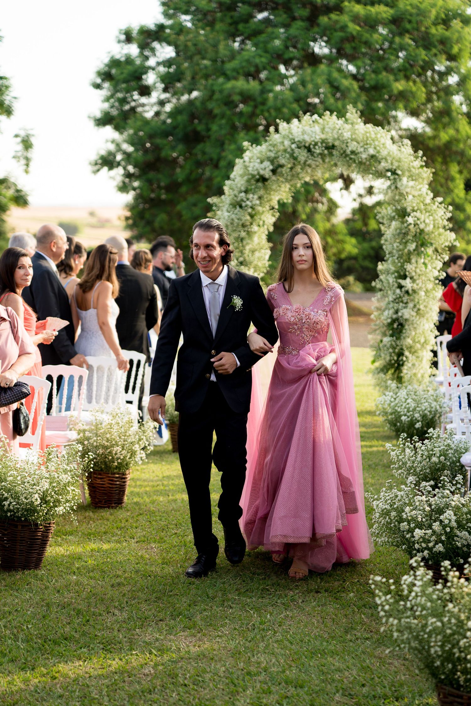Man in tuxedo escorts woman in pink gown down floral arch aisle at elegant outdoor wedding ceremony.