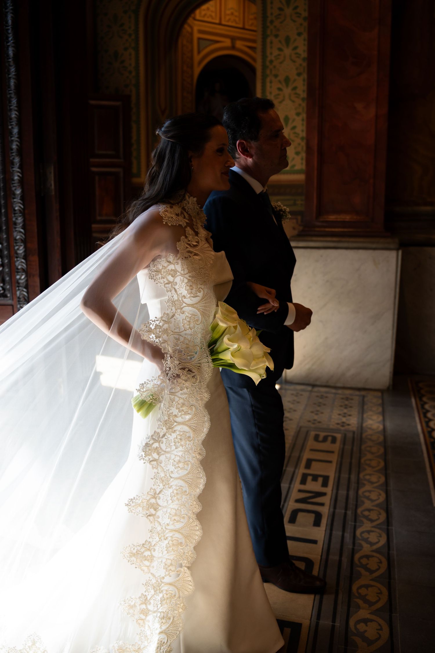 Bride in lace wedding gown with veil and bouquet stands beside groom in suit inside ornate marble building.