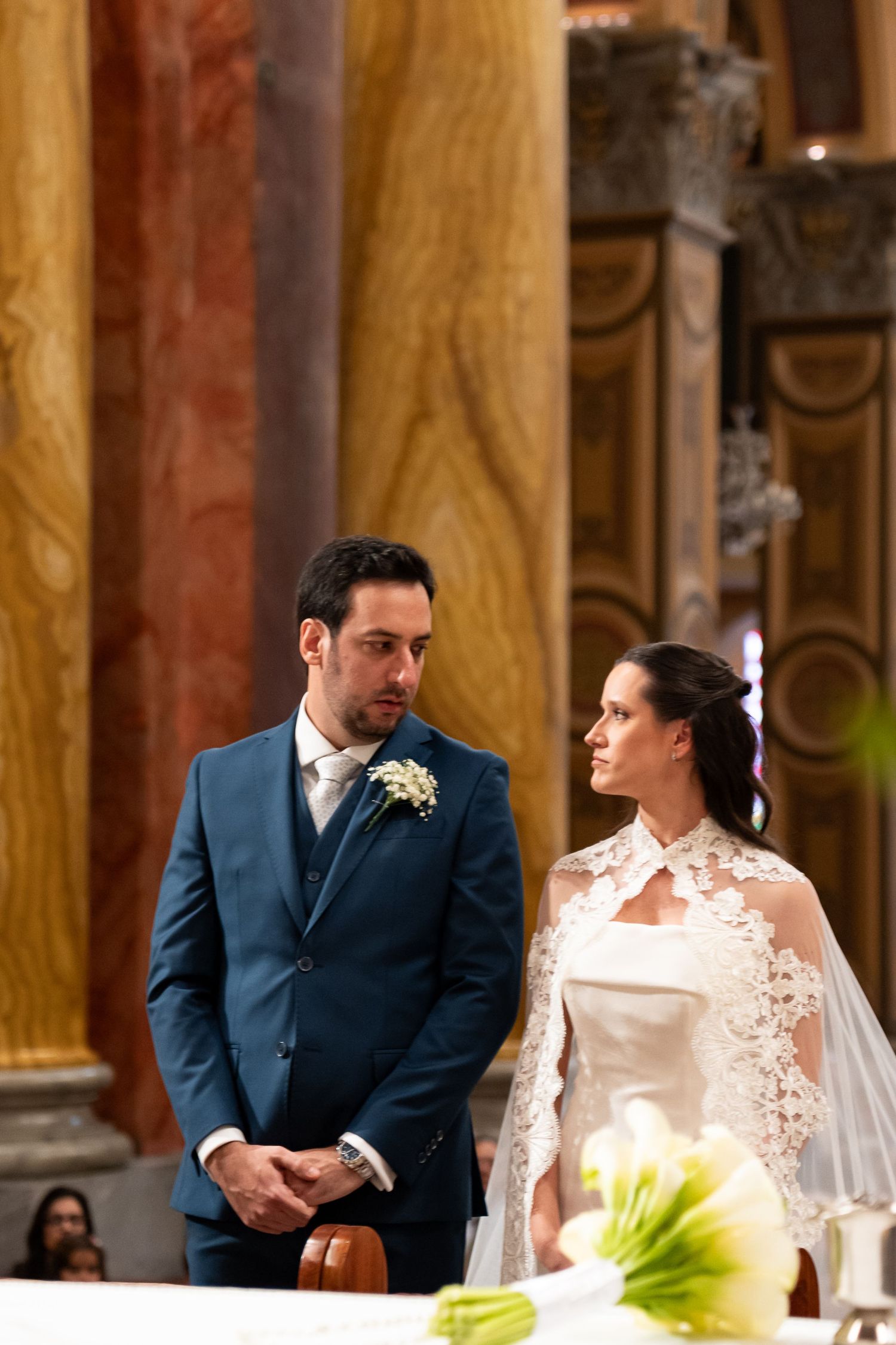 Bride in lace veil and groom in blue suit exchange vows at elegant church altar during wedding ceremony.