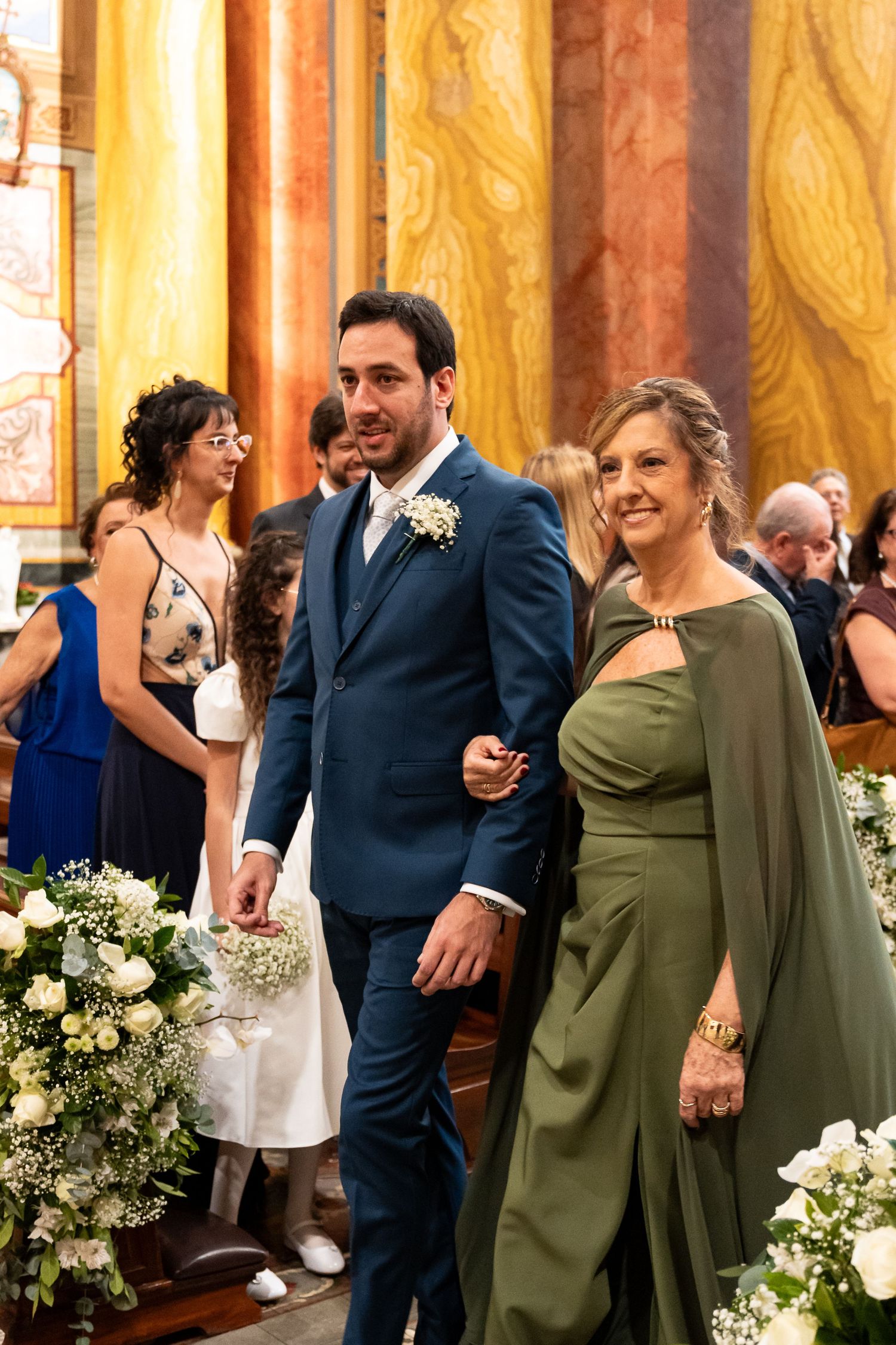 Groom in navy suit walks down church aisle with woman in green dress during wedding ceremony.