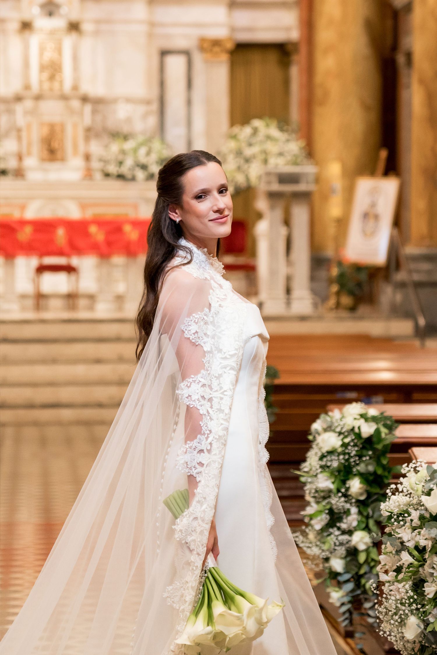 Elegant bride in lace wedding gown holding white calla lily bouquet inside ornate church.