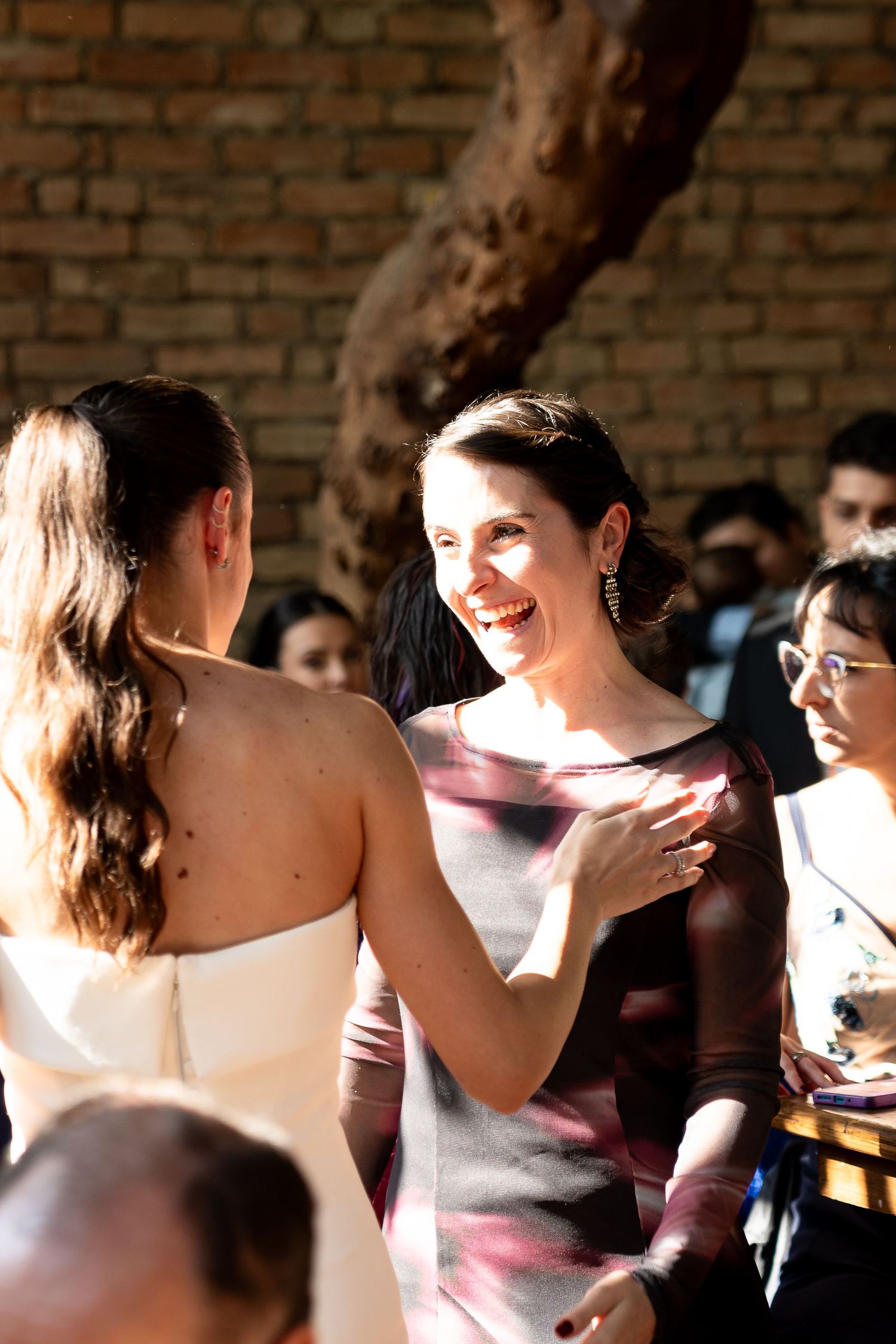 Two women sharing a joyful moment at a wedding, one in a white dress, the other smiling warmly in a gray gown.