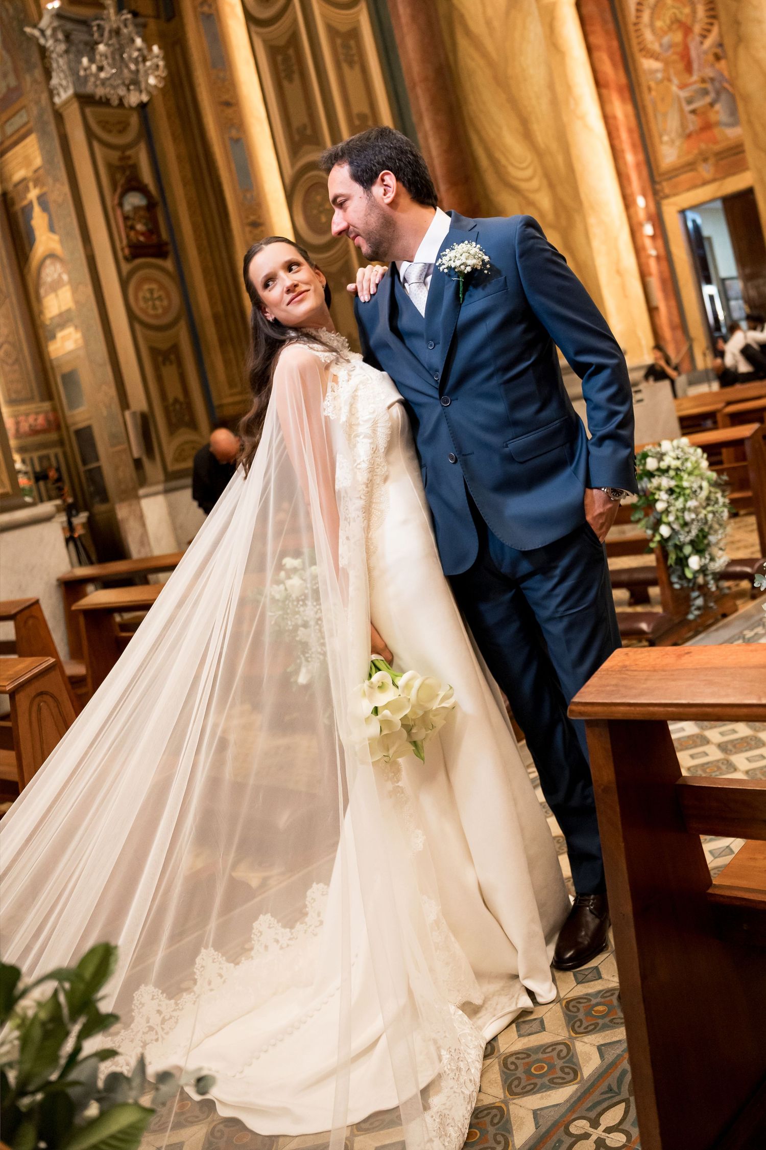 Bride in white gown and veil with groom in navy suit sharing a romantic moment inside an ornate church.