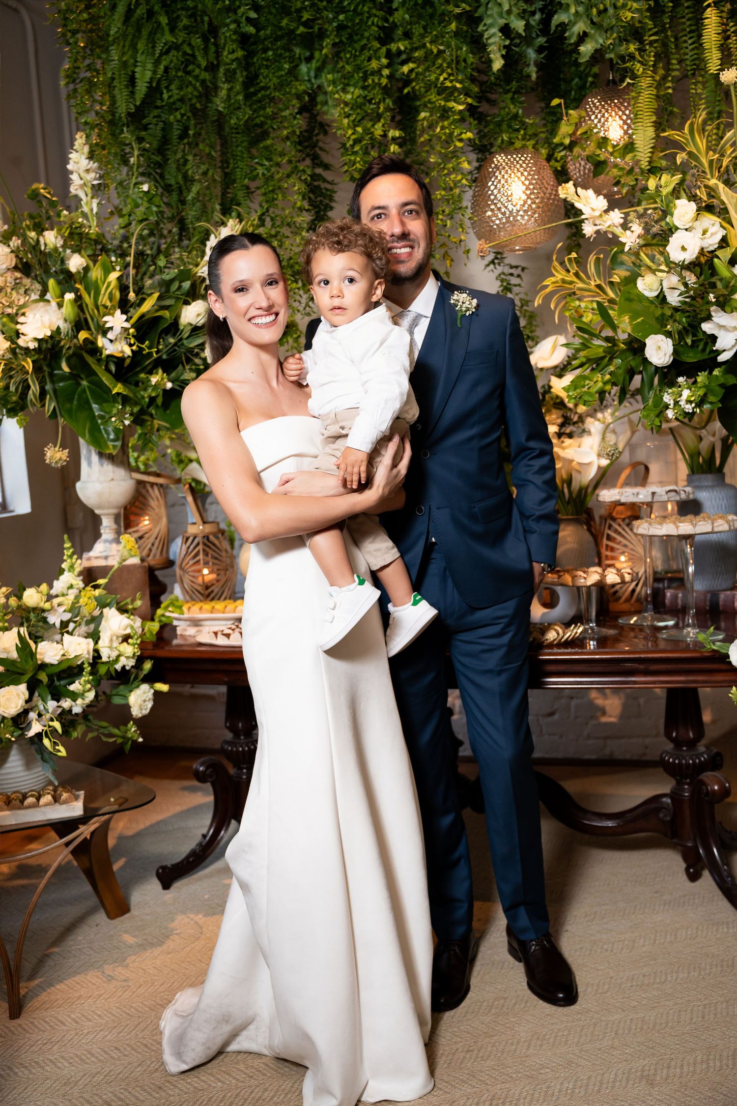 Bride in white gown and groom in navy suit pose with young child at elegant wedding reception.