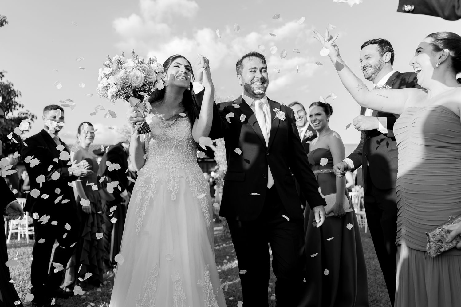 Black and white photo of a joyful bride and groom walking through confetti shower at outdoor wedding ceremony.