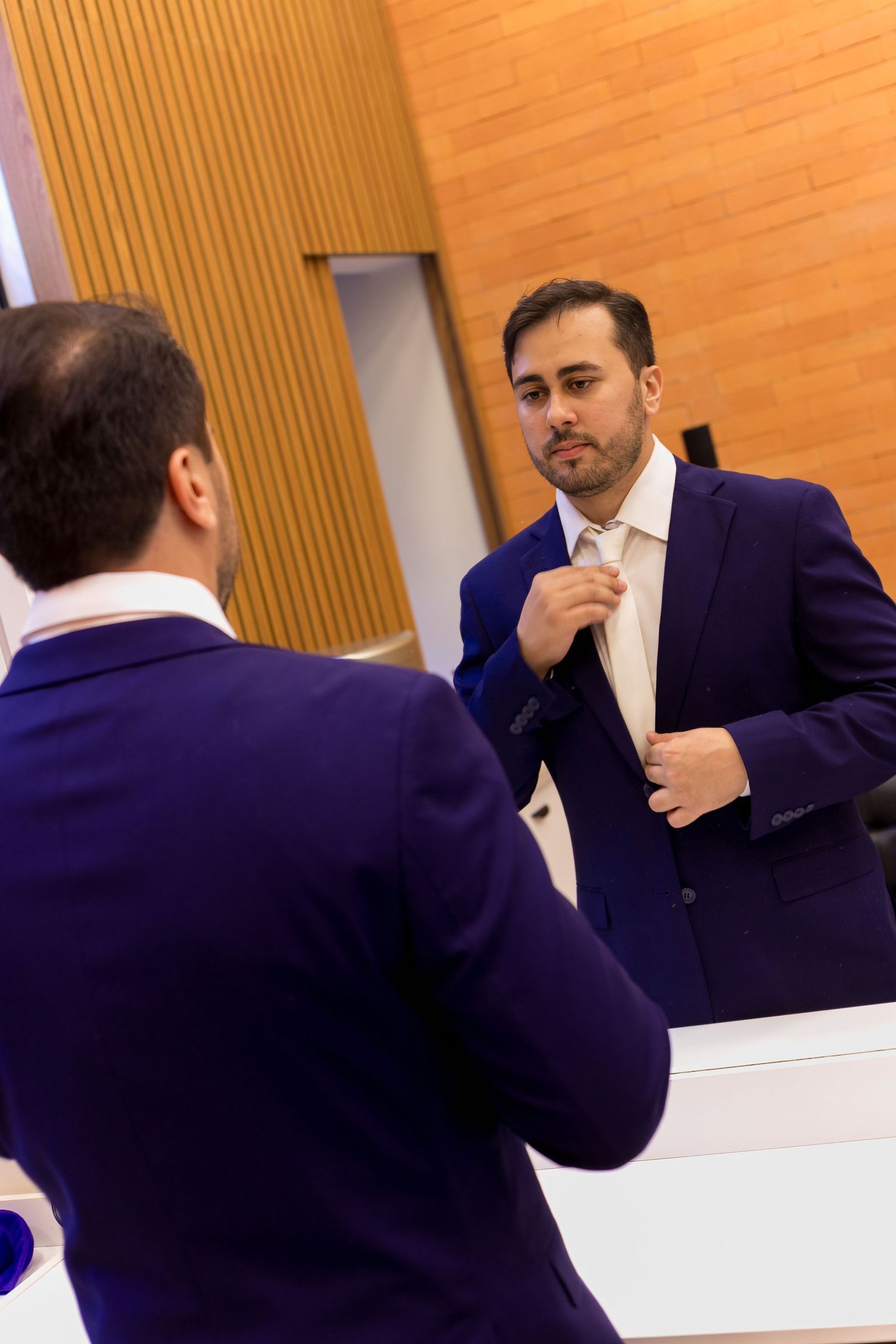 A man in a purple suit adjusts his tie while looking at his reflection in a large mirror.