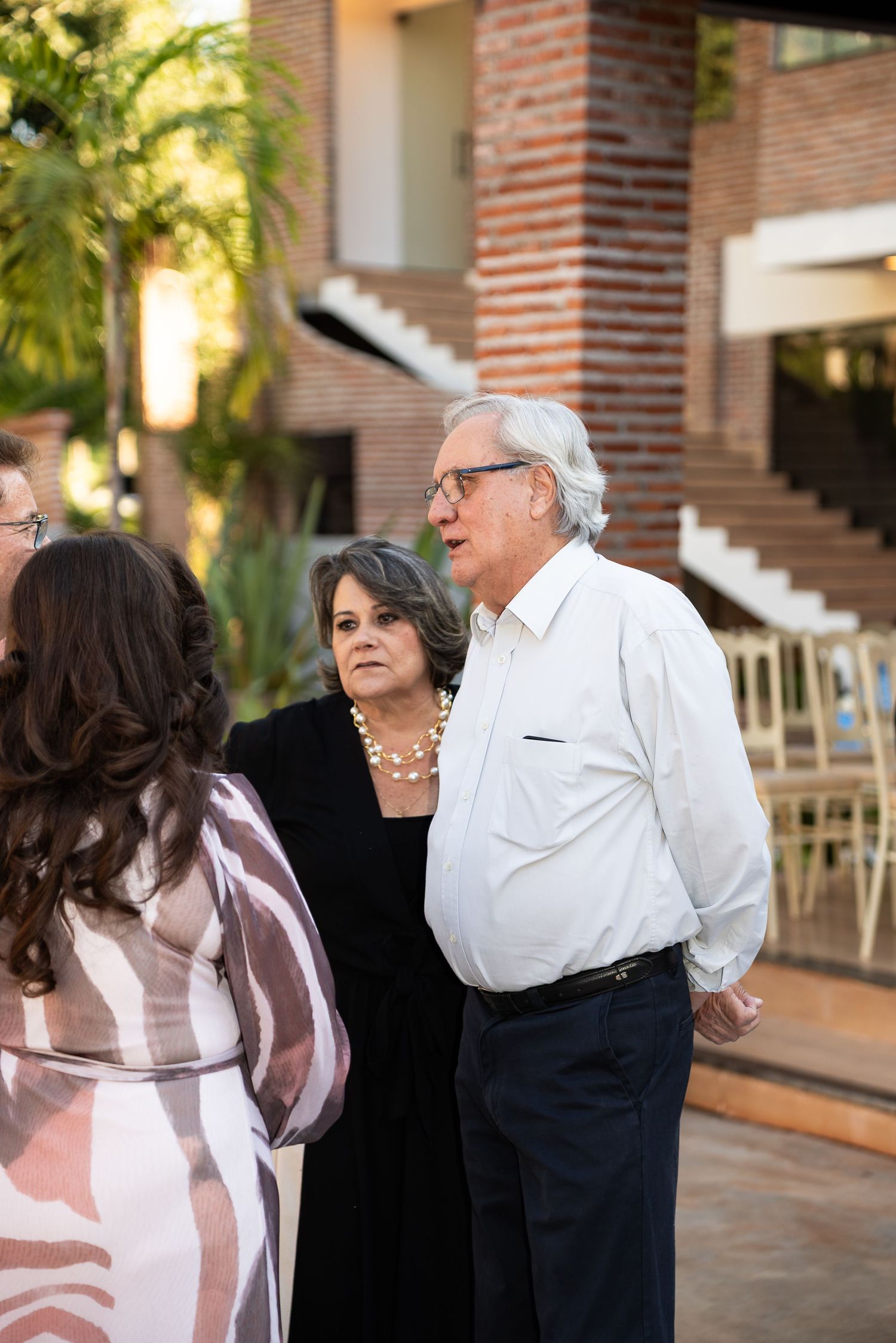 Elderly couple in formal attire conversing outdoors near a brick building with palm trees in background.
