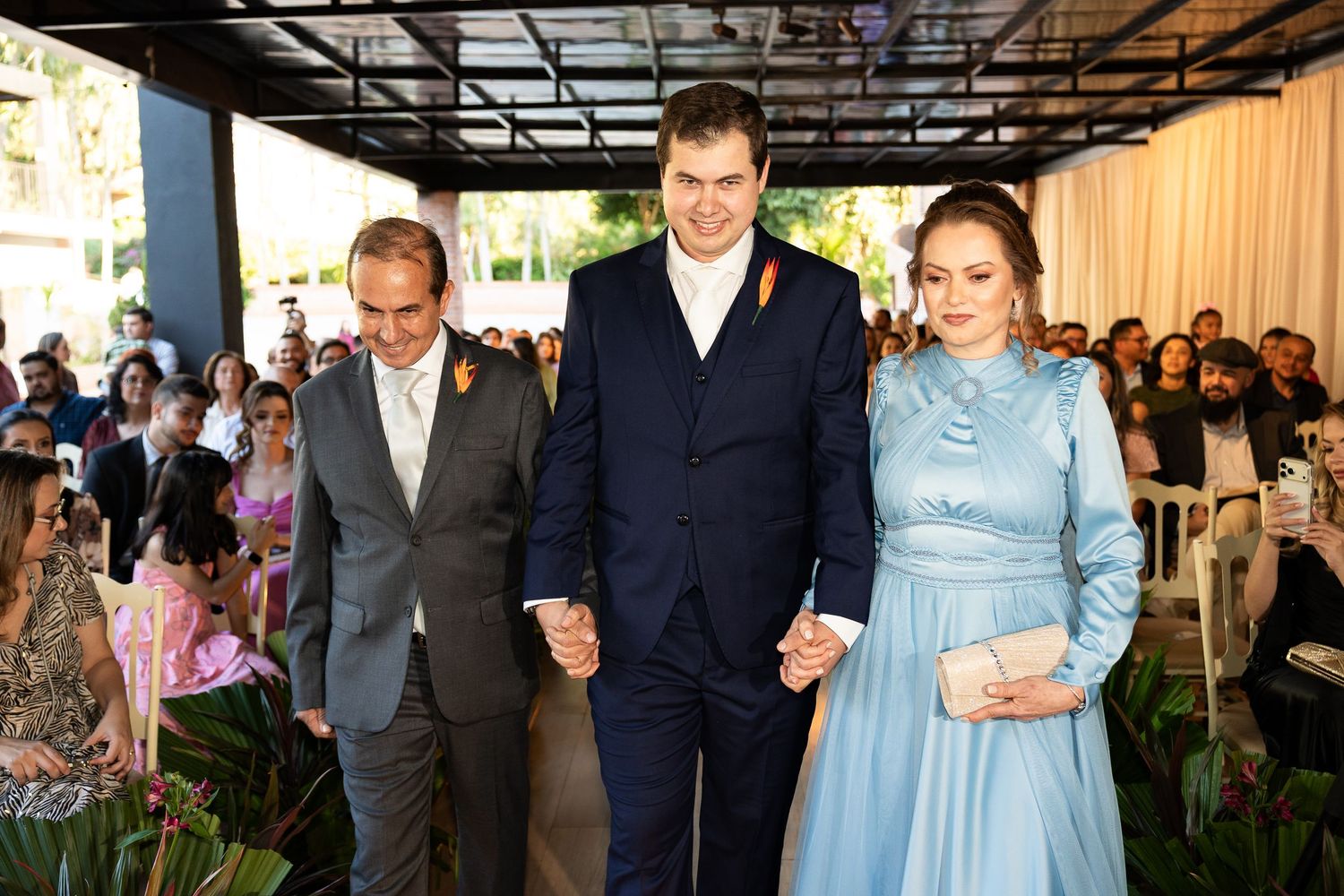 Two men in suits and a woman in a blue gown walk hand in hand at an elegant outdoor wedding ceremony.