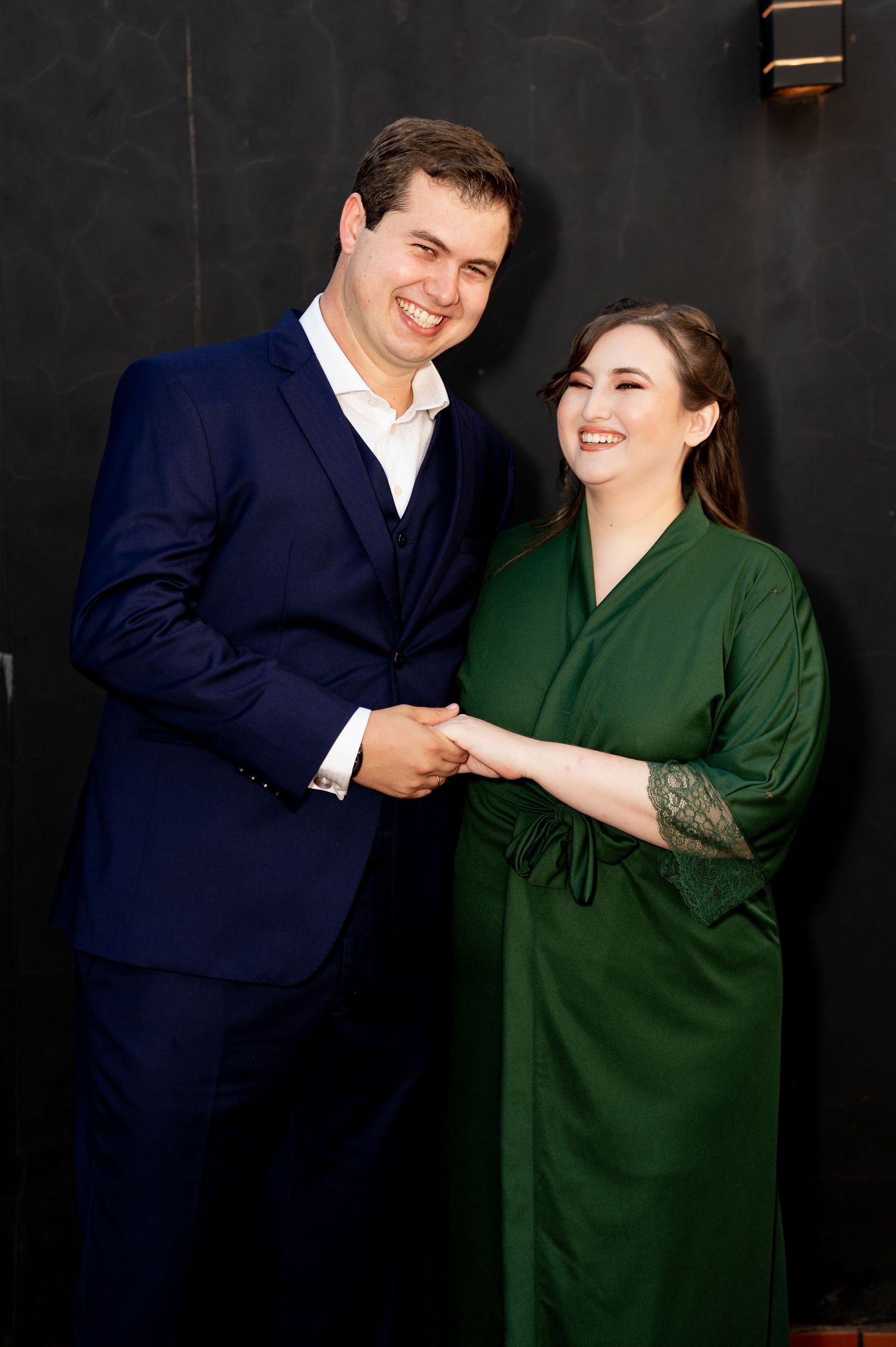 A smiling couple posing together, man in navy suit and woman in elegant green dress, holding hands against dark background.