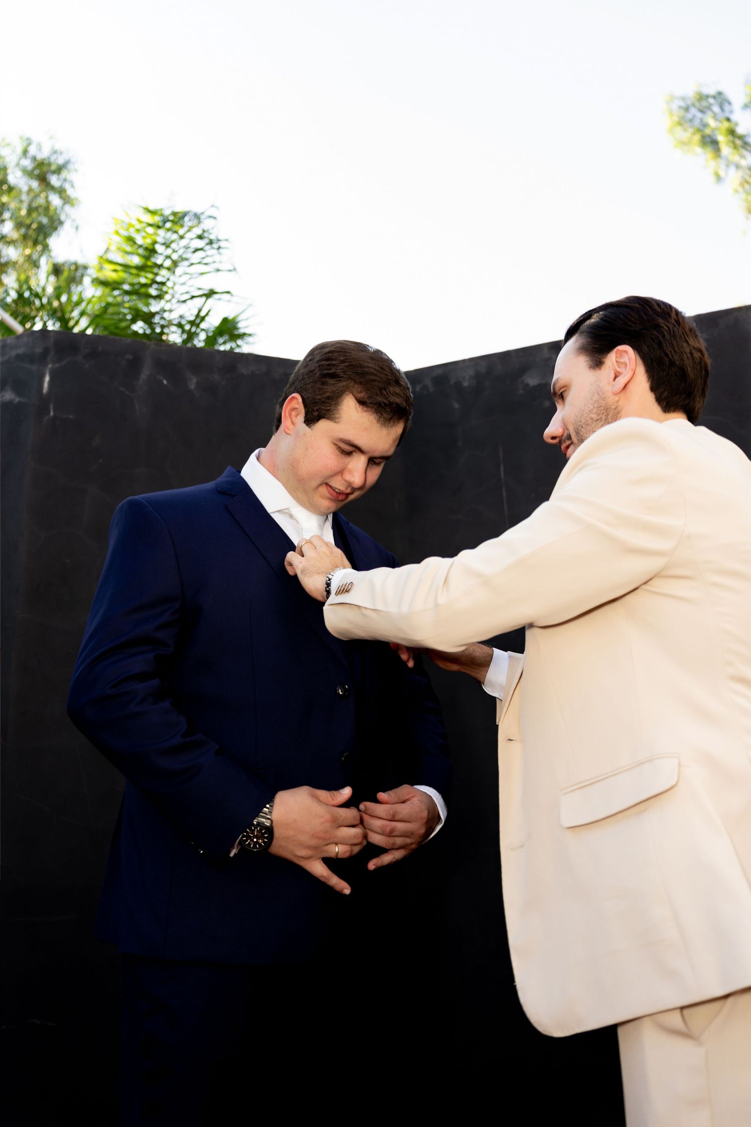 Two men in suits, one in navy and one in cream, adjusting a boutonniere outdoors before a wedding ceremony.