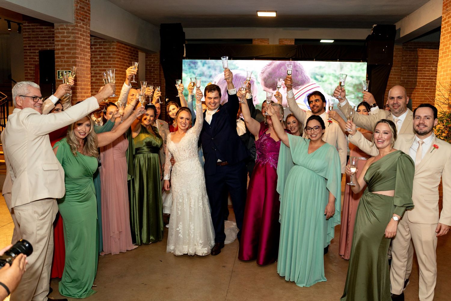 Wedding party celebrating with raised glasses, bride in white gown, bridesmaids in green and pink dresses, indoor venue.