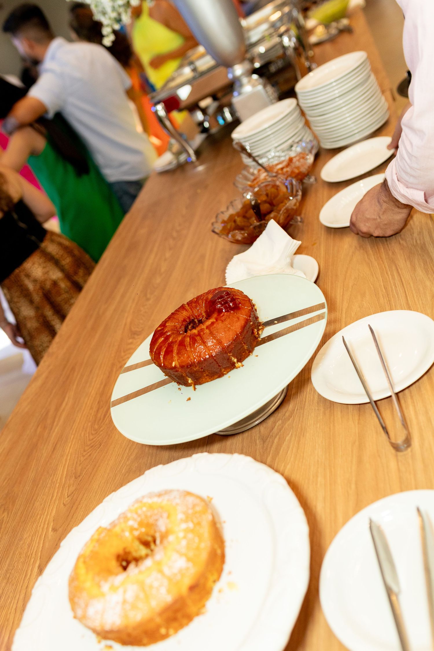 A buffet table displays glazed donuts and pastries on white plates with tongs, as guests and stacked dishes appear in the background.