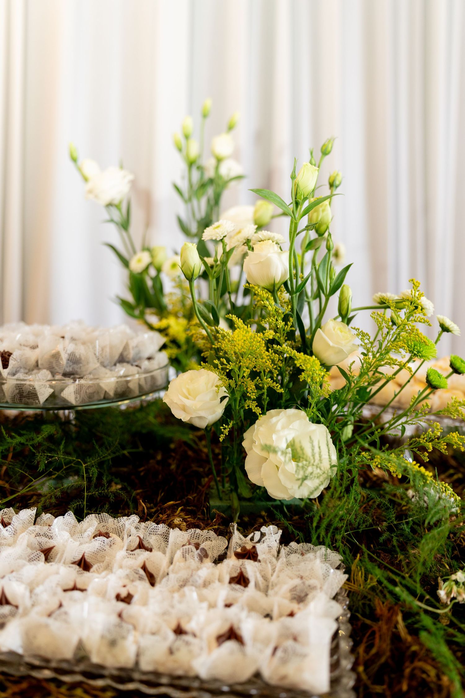 Elegant wedding dessert table with white roses, yellow flowers, and wrapped chocolates on a moss-covered surface.