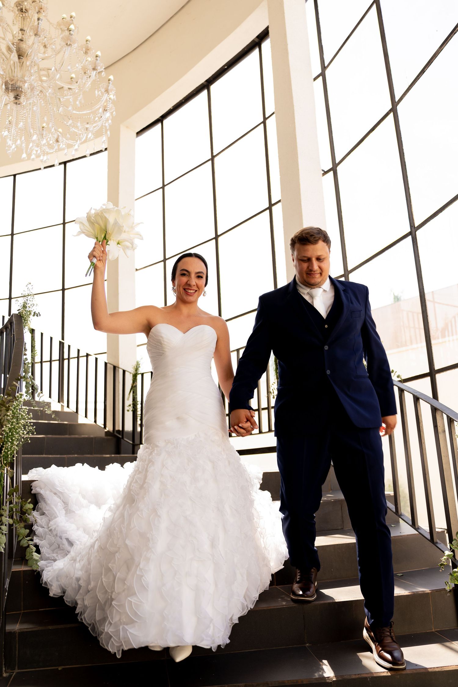 Bride in white gown holding bouquet and groom in navy suit hold hands descending elegant staircase.