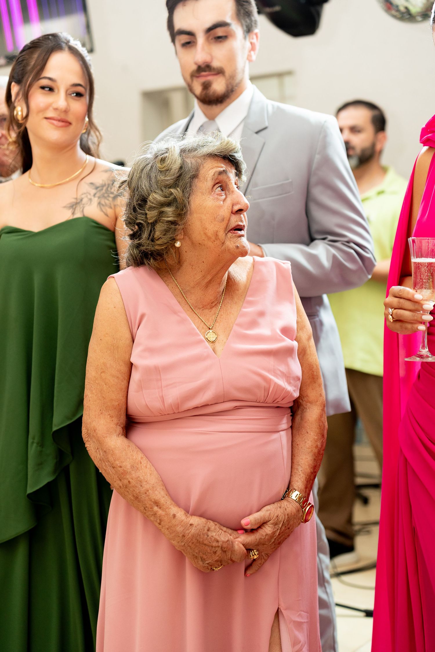 Elderly woman in pink dress at formal event, surrounded by guests in elegant attire, looking up with expressive eyes.