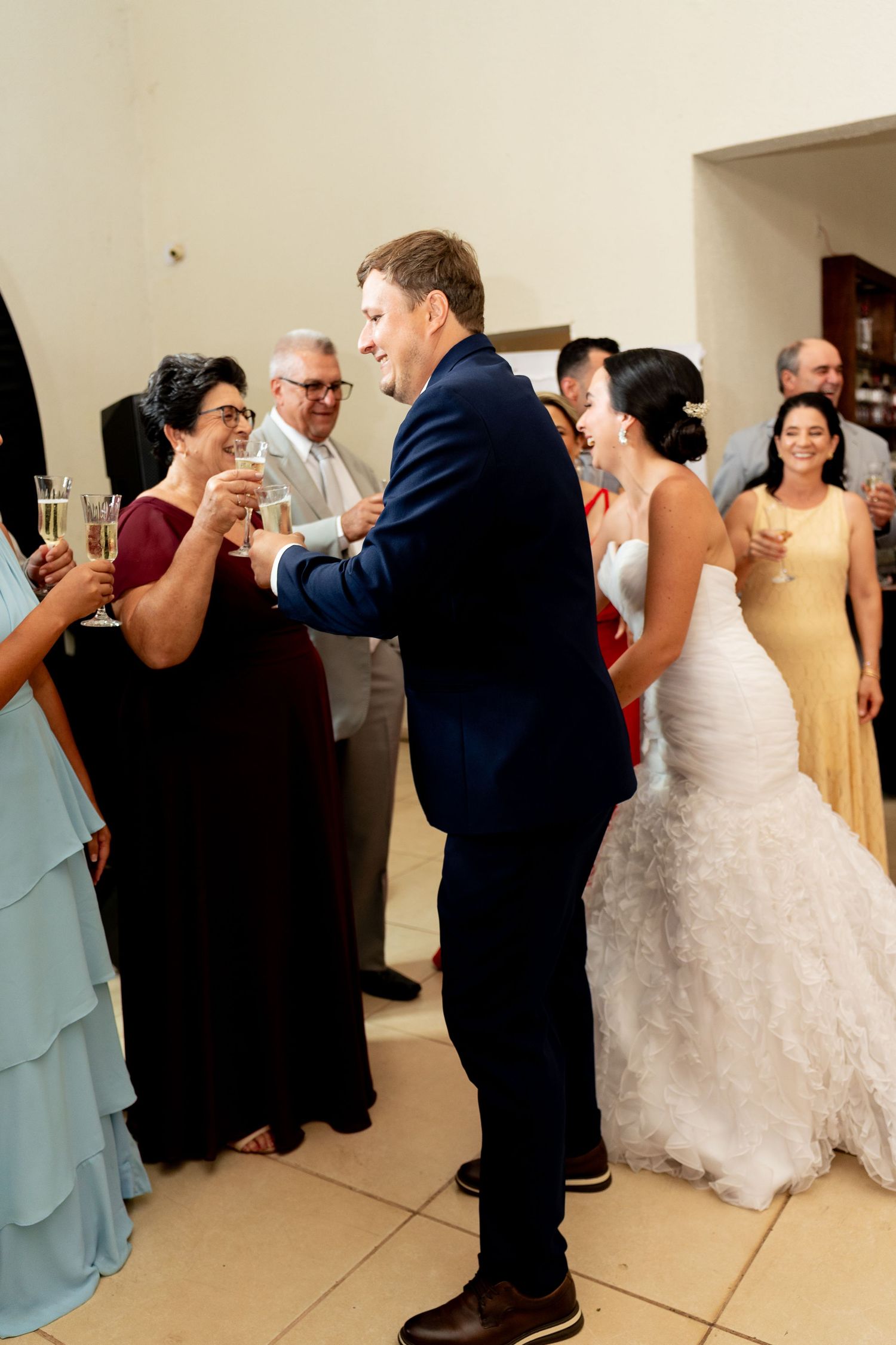 Bride in white gown and groom in navy suit toasting champagne with guests at elegant wedding reception.