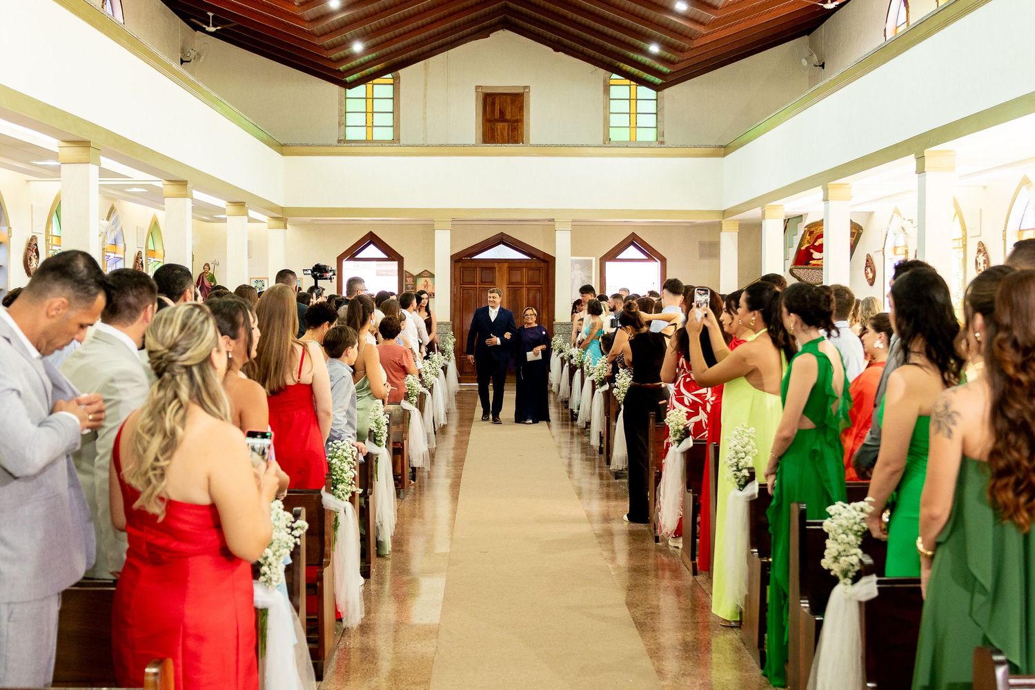 Wedding ceremony in a bright church with guests in vibrant dresses lining the decorated aisle.