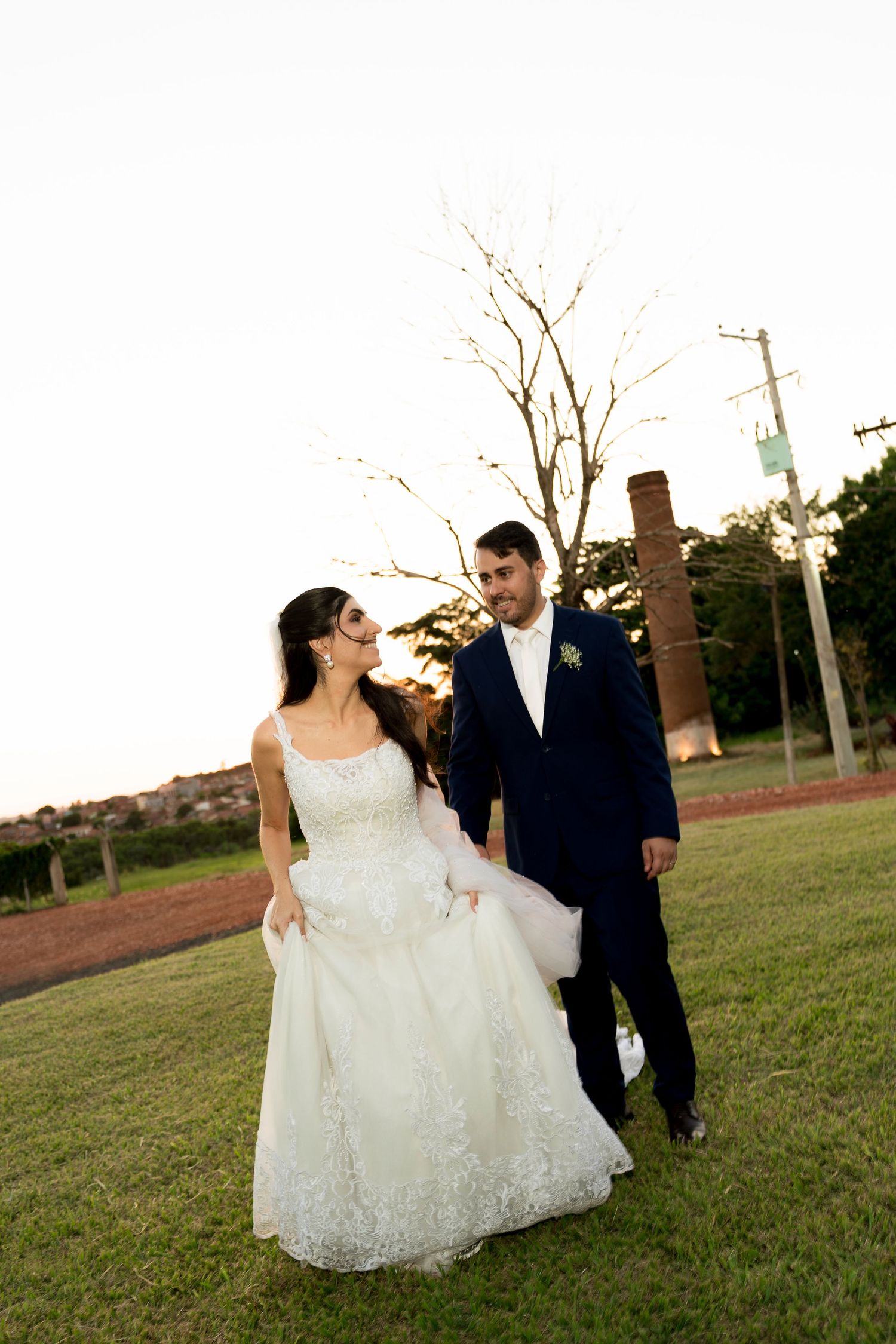 Bride in white lace gown and groom in navy suit walk hand in hand on green lawn at outdoor wedding.