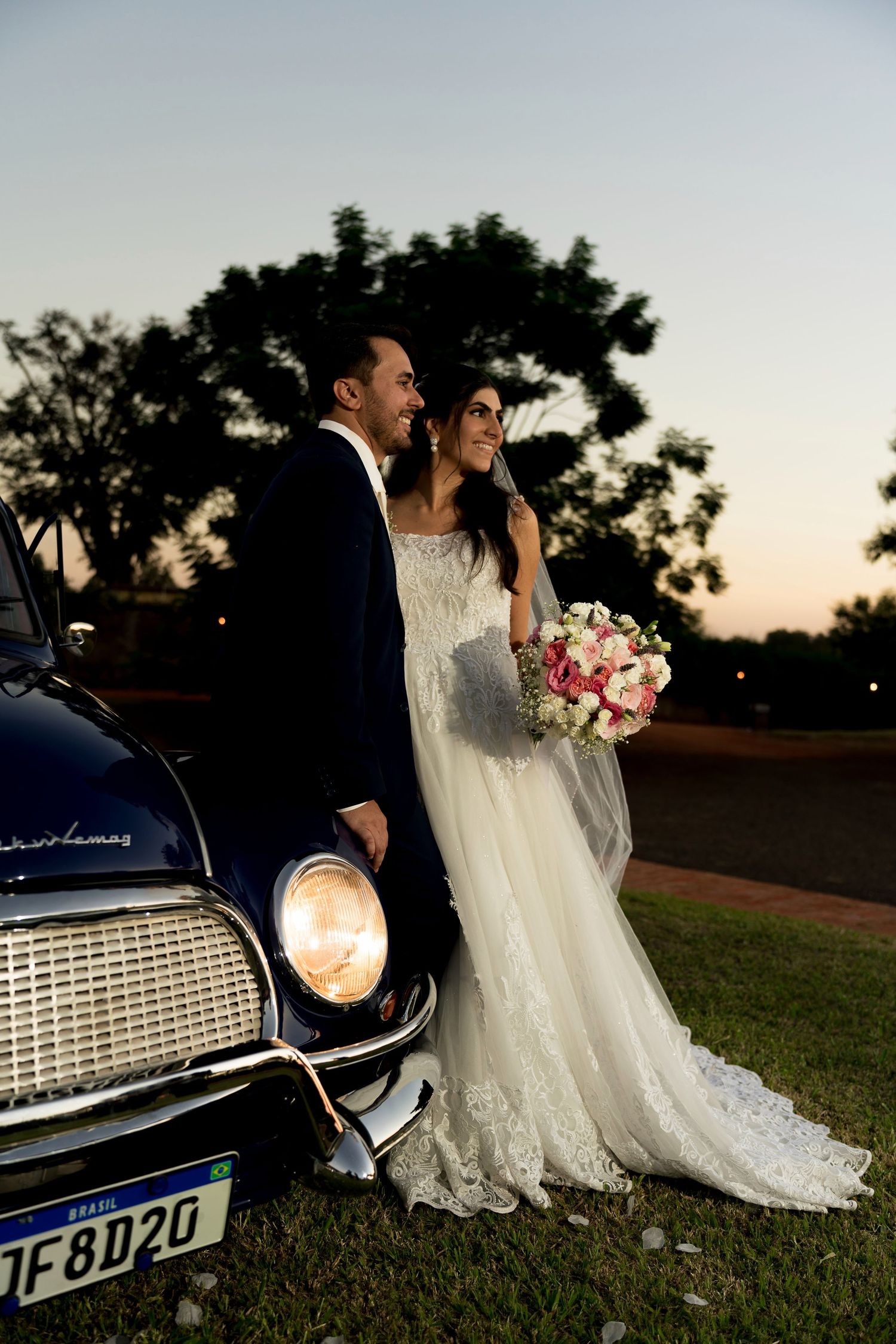 Bride and groom pose beside a vintage classic car at dusk, bride holding a pink and white bouquet in a lace wedding gown.
