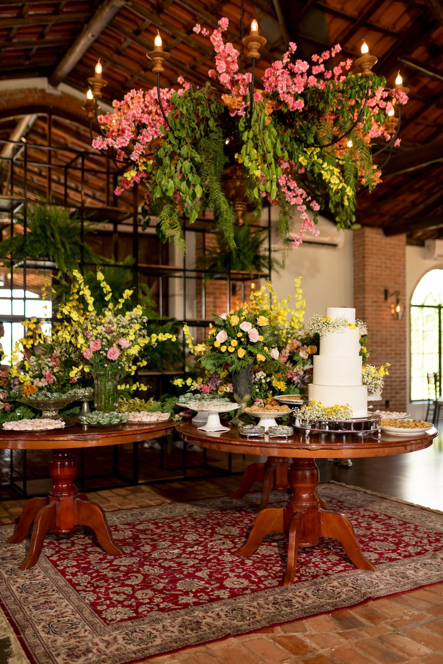 Elegant wedding dessert table with tiered cake, colorful floral arrangements, and hanging pink blooms in a rustic venue.
