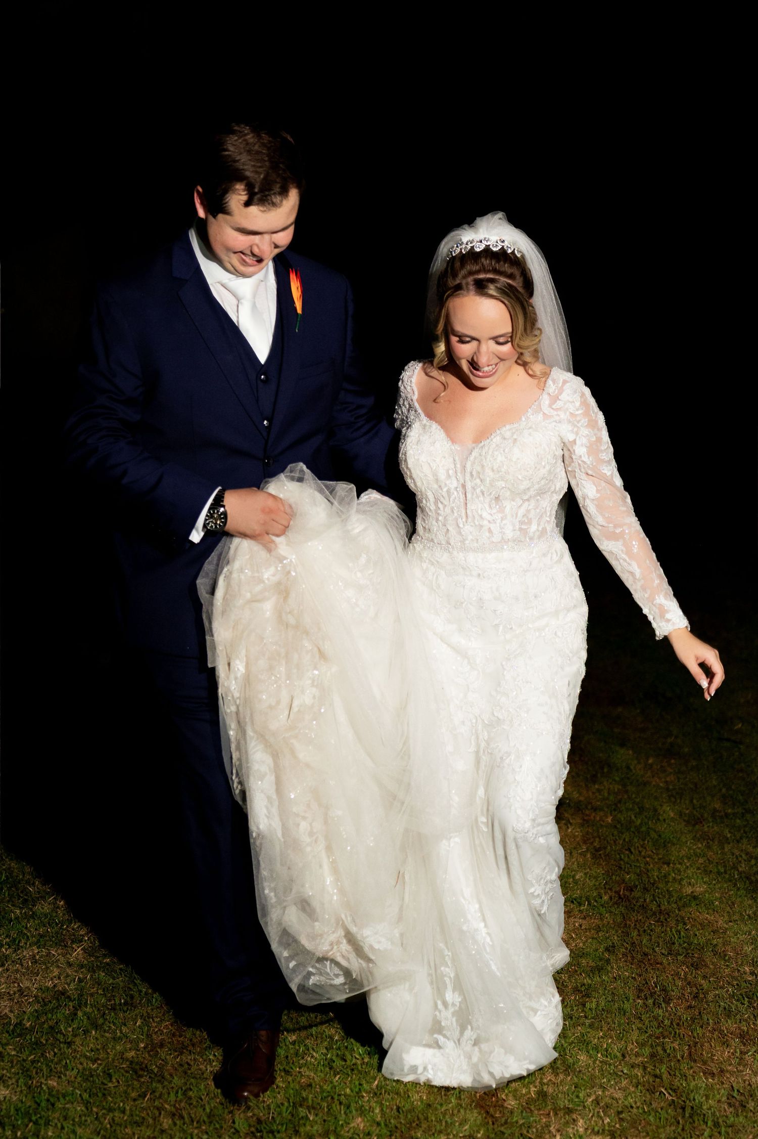 A smiling bride in a lace gown and veil walks with her groom in a navy suit on their wedding night.