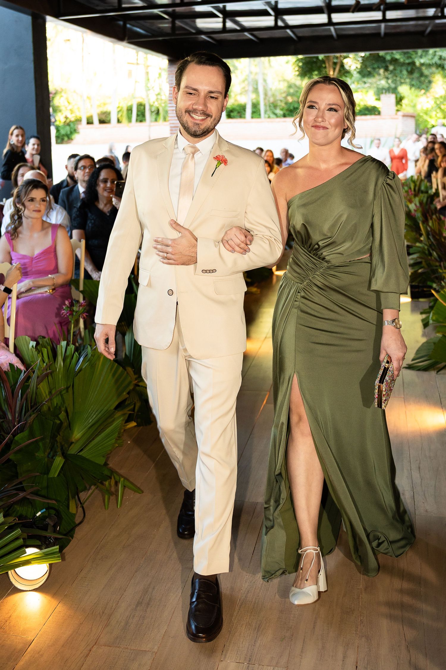 Elegant couple walking at a wedding, man in beige suit, woman in olive green one-shoulder gown with slit.