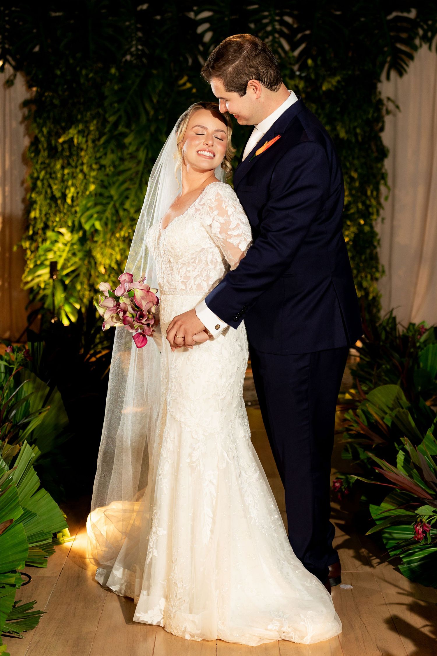 Bride in lace gown and veil holds bouquet while groom in navy suit embraces her before tropical greenery backdrop.