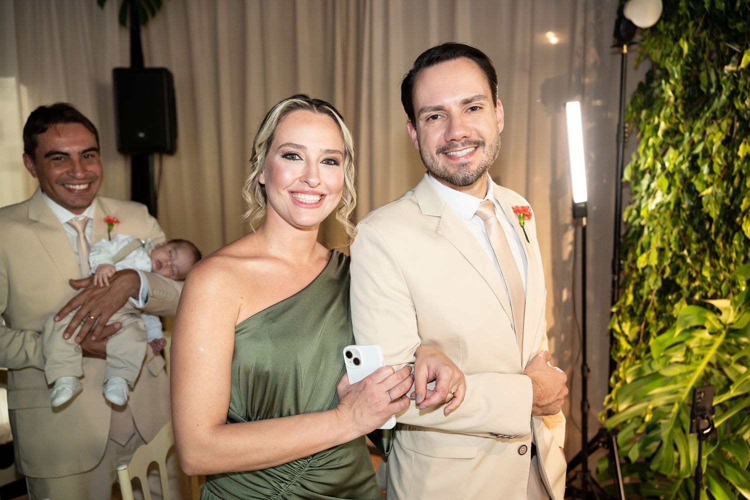 Couple smiling at wedding reception, woman in green dress, man in beige suit, greenery backdrop.
