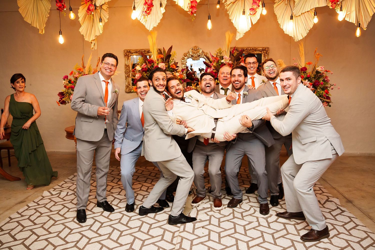 Groomsmen in gray suits joyfully lift the groom at a festive wedding reception with floral decor.