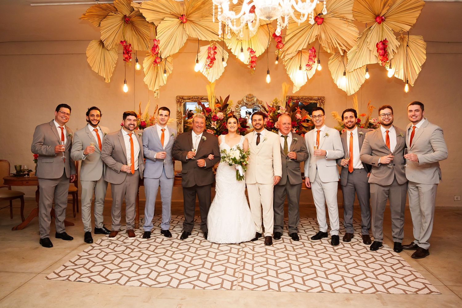 Wedding party poses together, bride and groom centered among groomsmen in gray suits with orange ties under floral decor.
