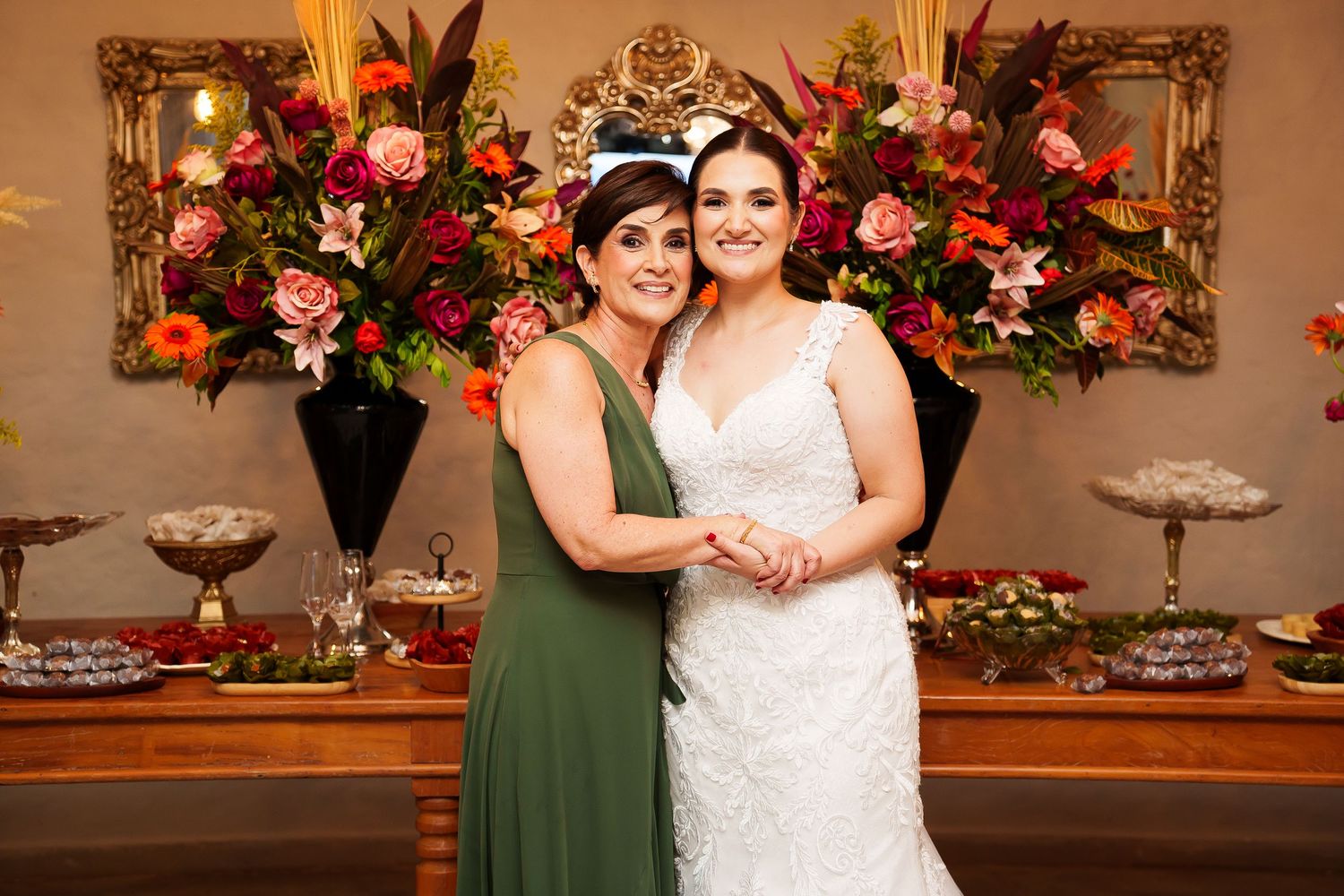 Bride in white lace gown poses with woman in green dress before floral backdrop at wedding reception.