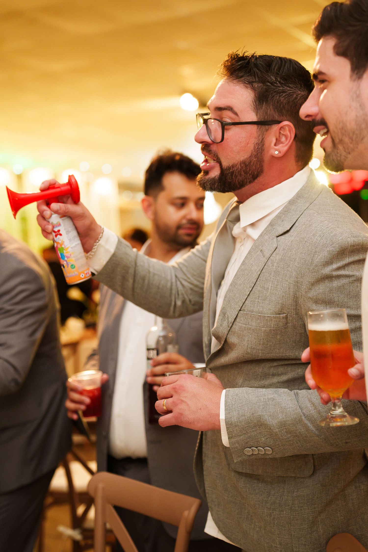 Man in gray suit blowing air horn while holding beer glass at indoor party celebration event.