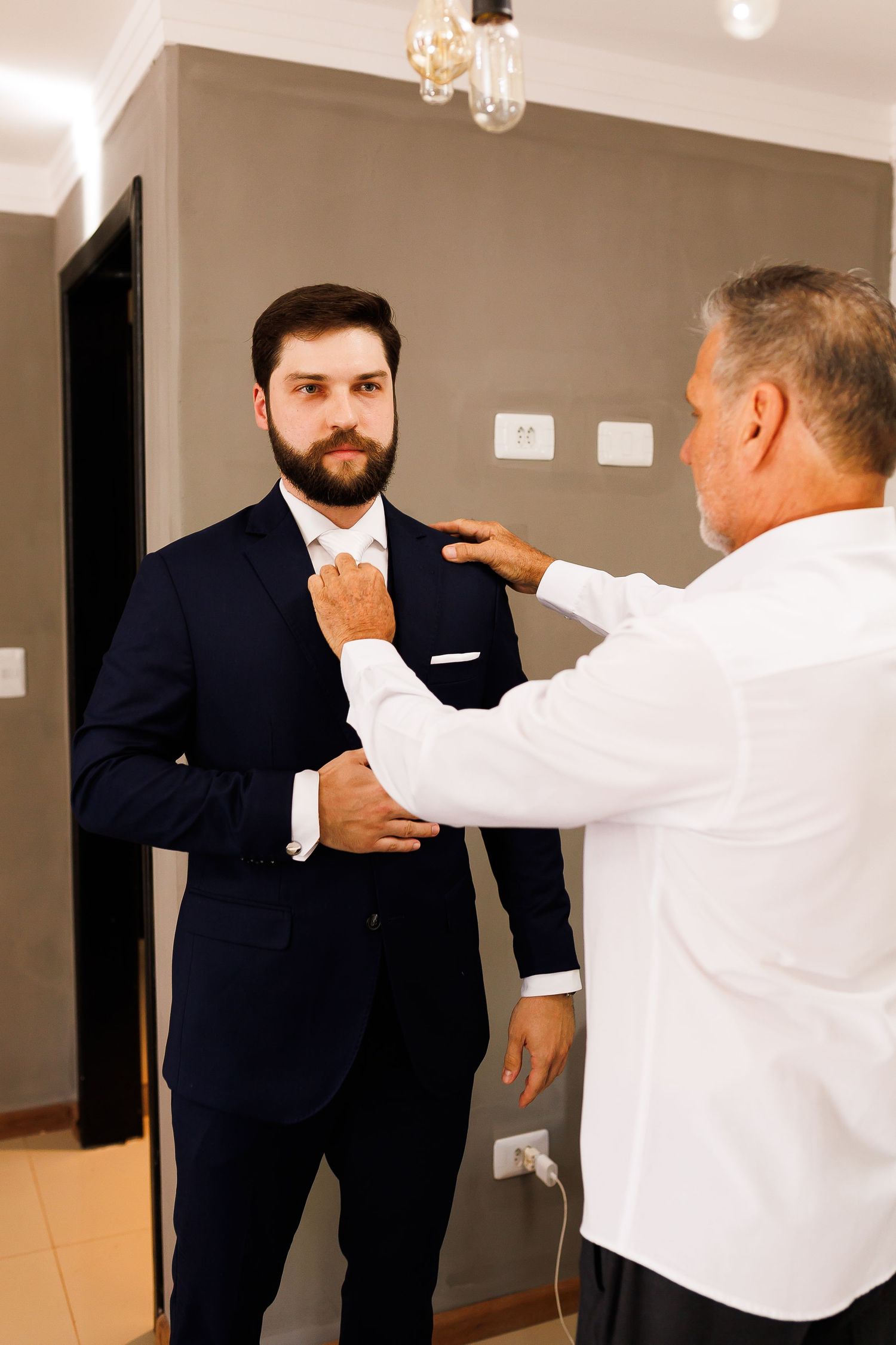 An older man in a white shirt adjusts the tie and jacket of a bearded groom in a navy suit before a wedding.