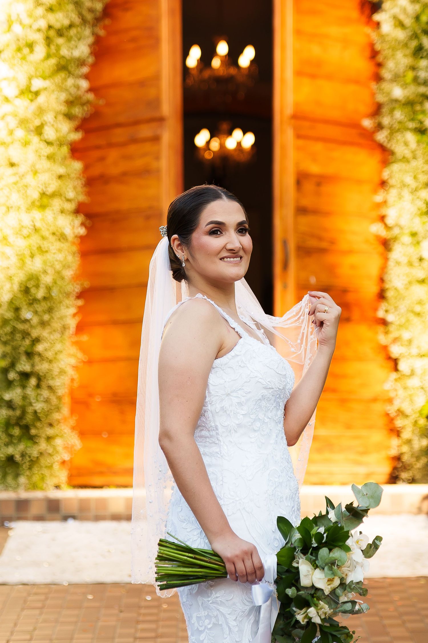 Smiling bride in white lace gown and veil holds bouquet of white flowers before ornate wooden doors.