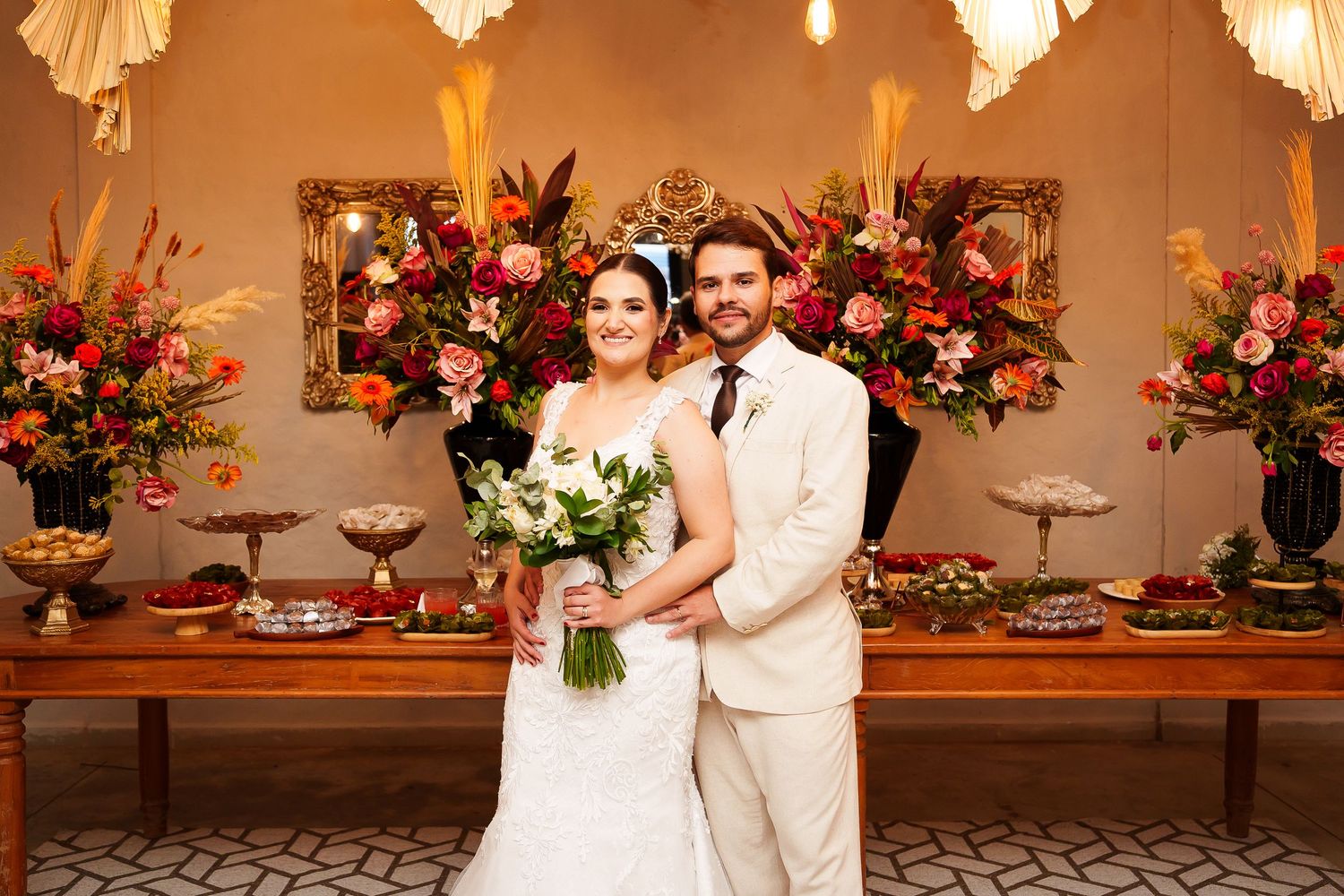 Bride and groom pose together in front of a floral-decorated wedding reception table with vibrant red and orange arrangements.