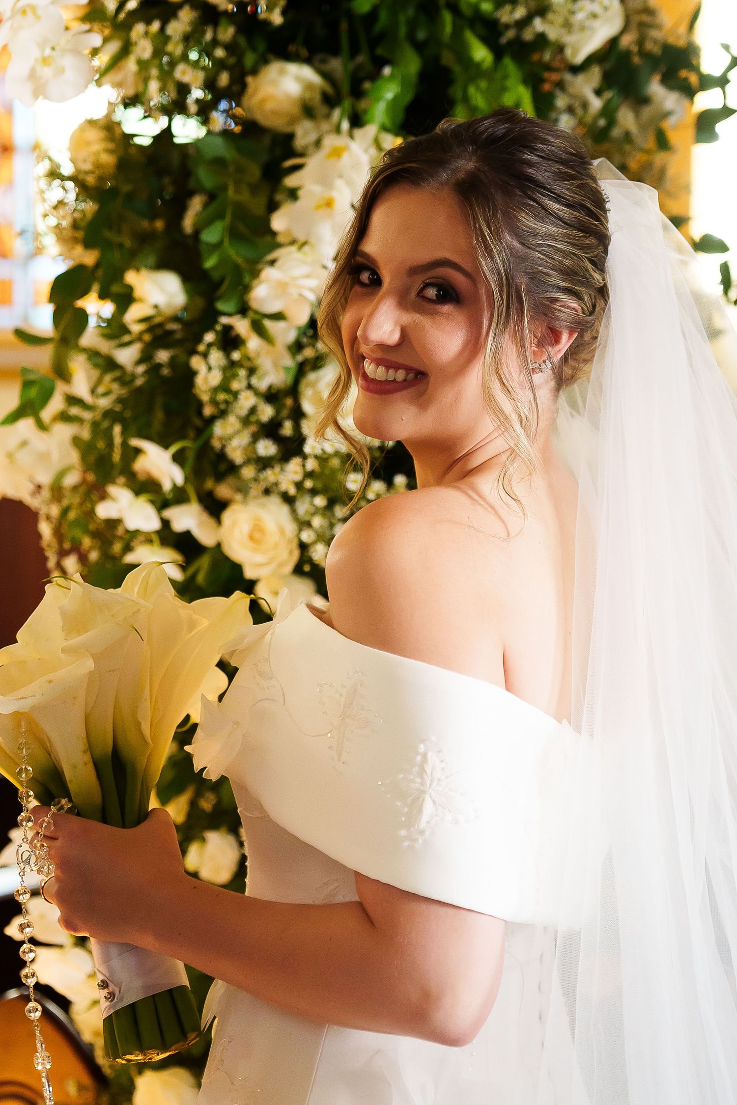 Smiling bride in off-shoulder white gown and veil holds yellow lilies near lush white floral backdrop.