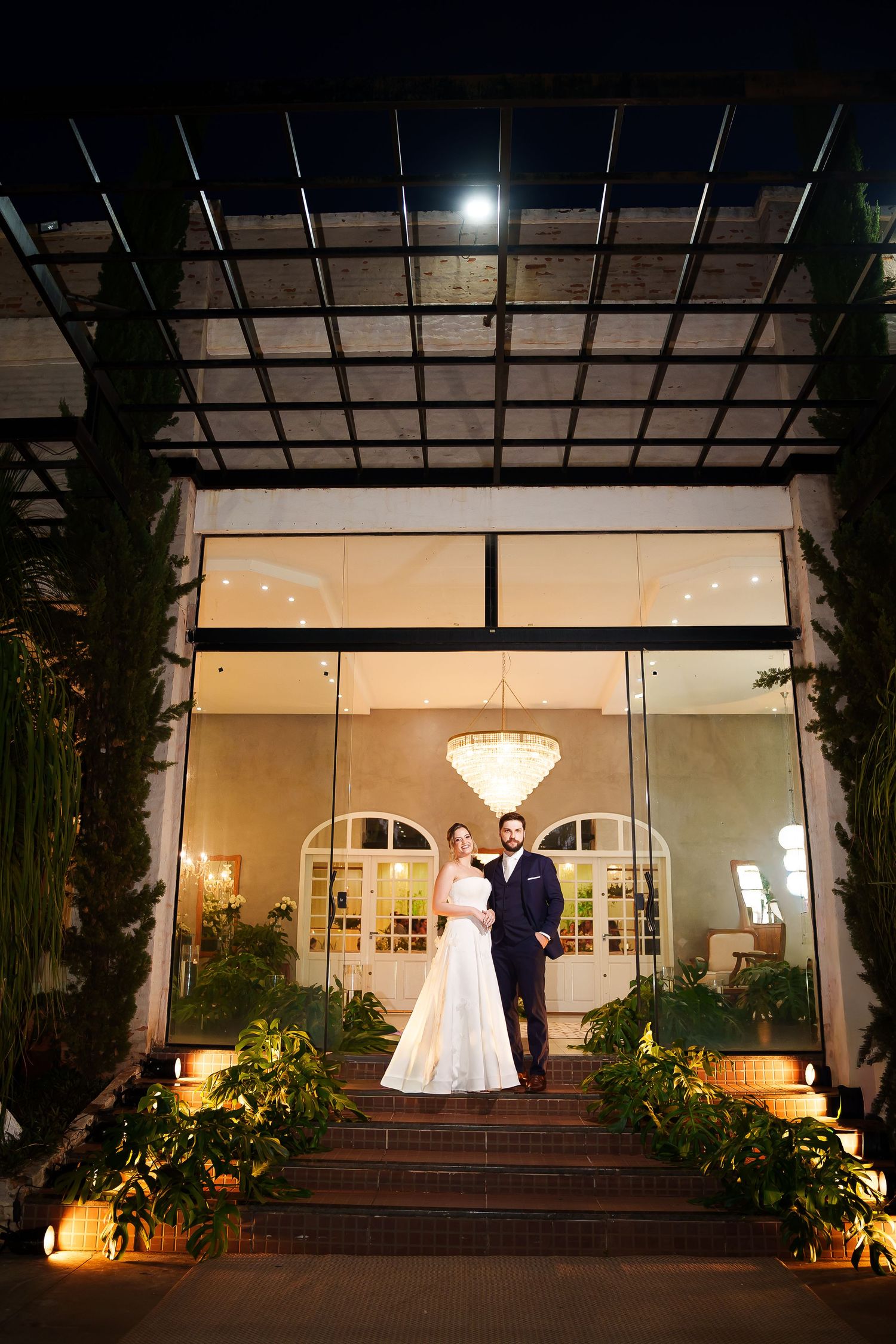 Bride and groom pose on steps outside an elegant venue entrance at night, with a chandelier glowing behind them.