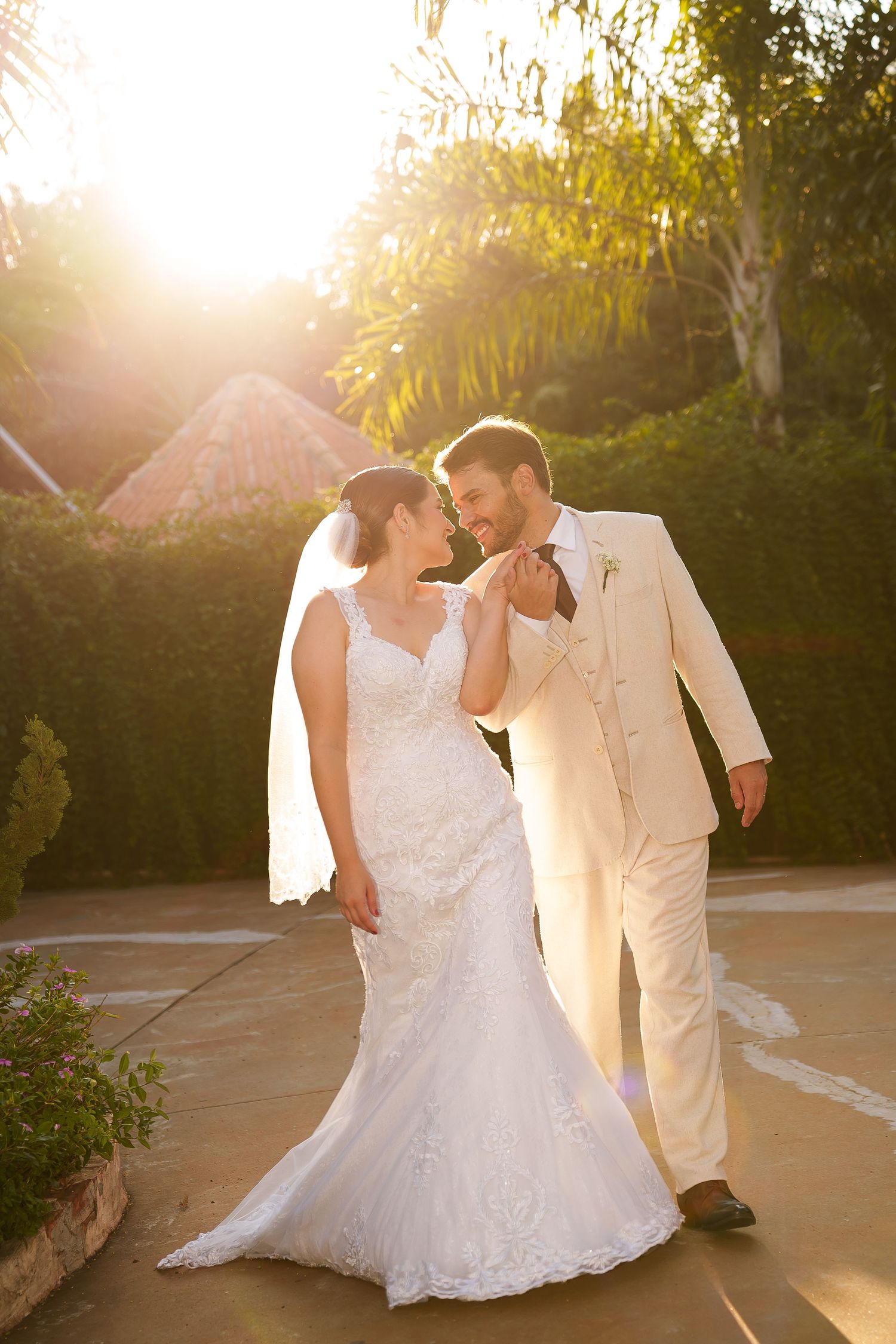 Bride in lace gown and groom in ivory suit share a romantic moment outdoors in golden sunset light.