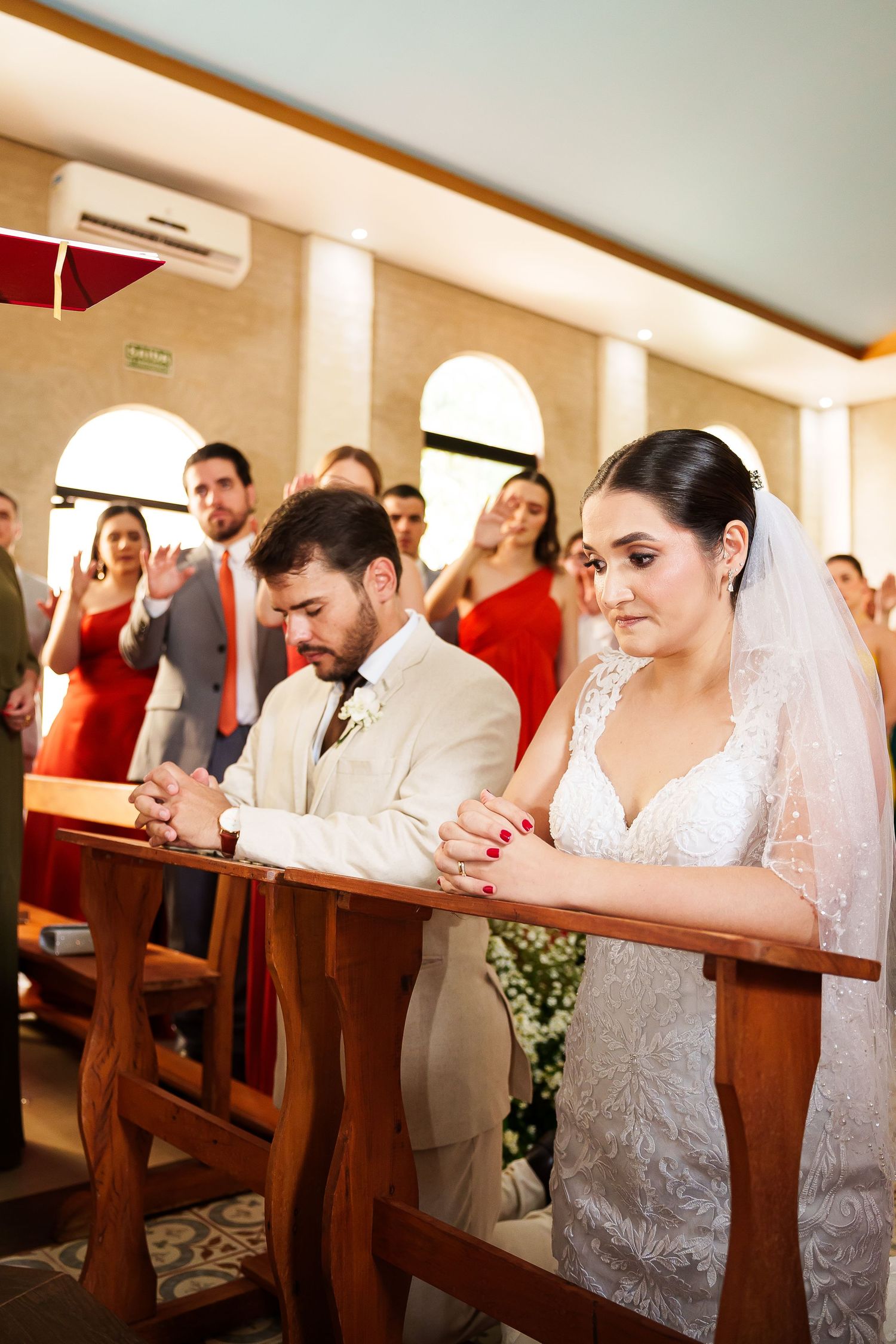 Bride and groom kneeling in prayer at church altar during wedding ceremony, guests in background.