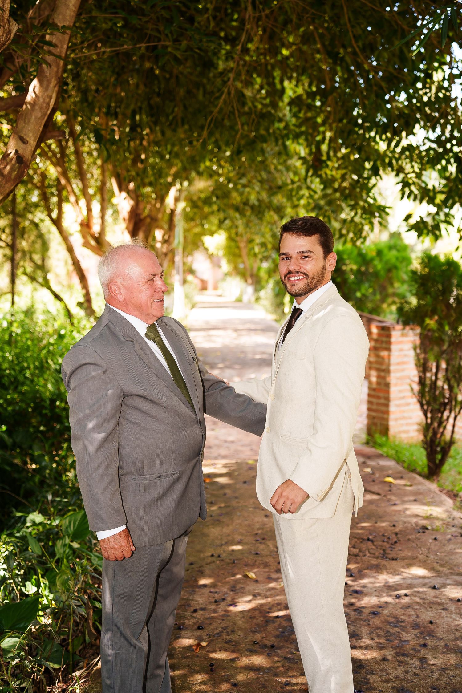 Two men in suits stand on a tree-lined path, smiling, likely at a wedding celebration outdoors.