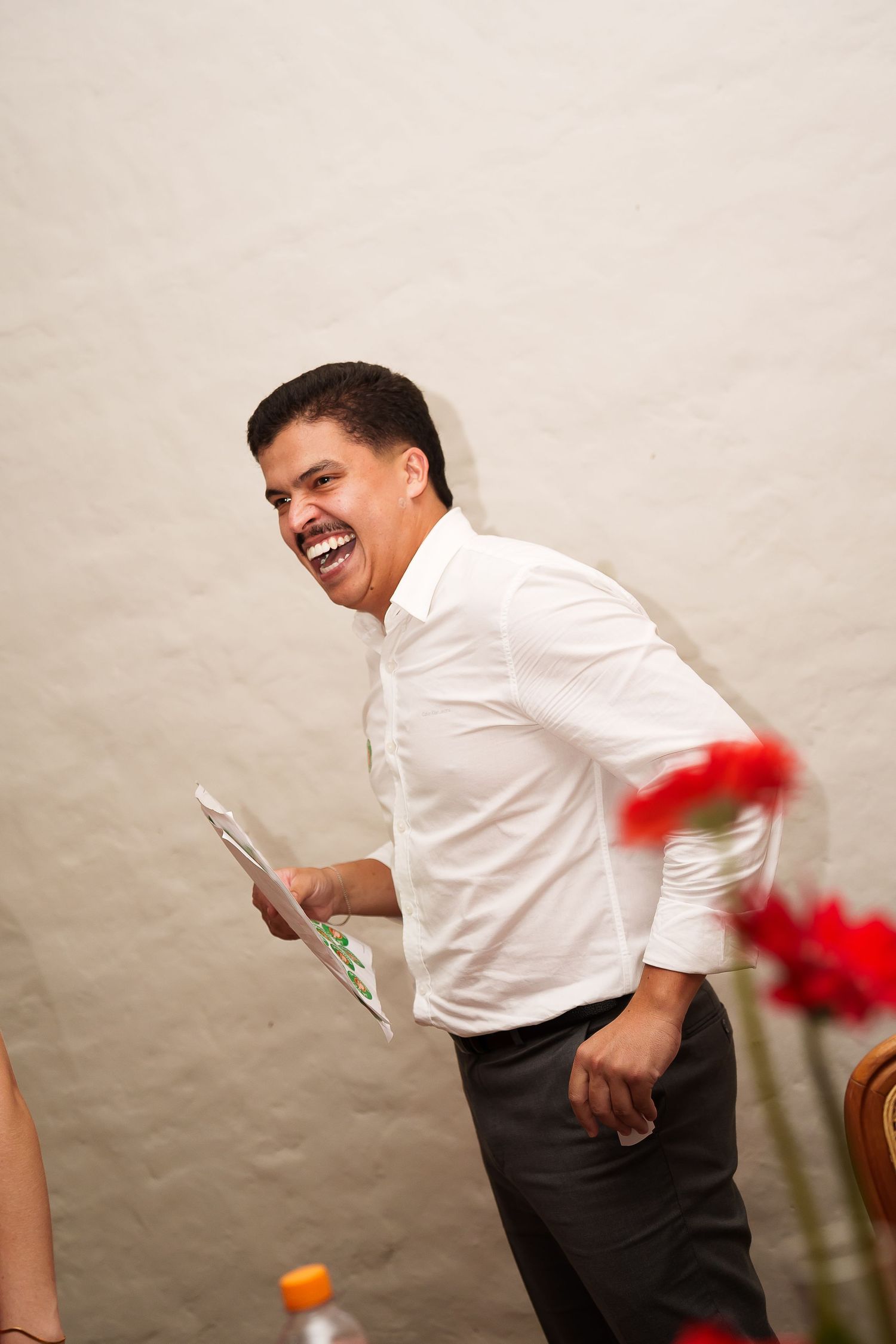 A laughing man in a white shirt holds papers at an indoor event, with red flowers visible in the background.