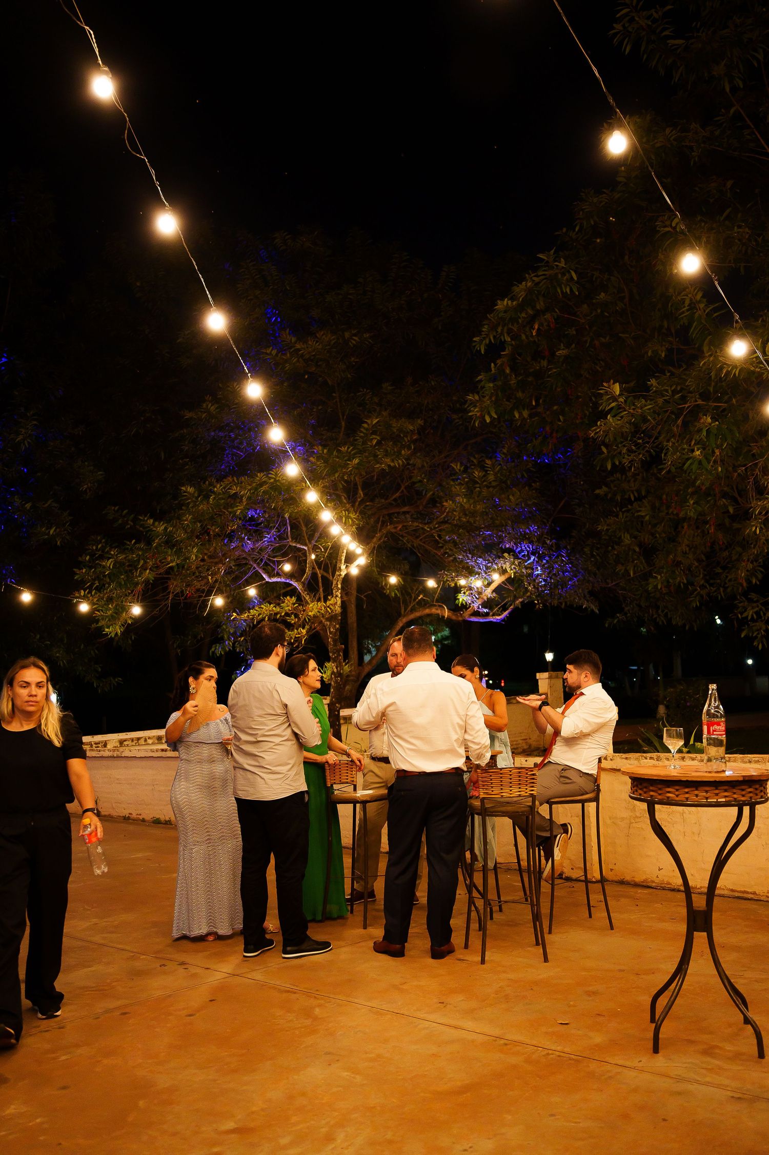 Outdoor evening party with string lights, guests mingling around high tables, surrounded by trees at night.