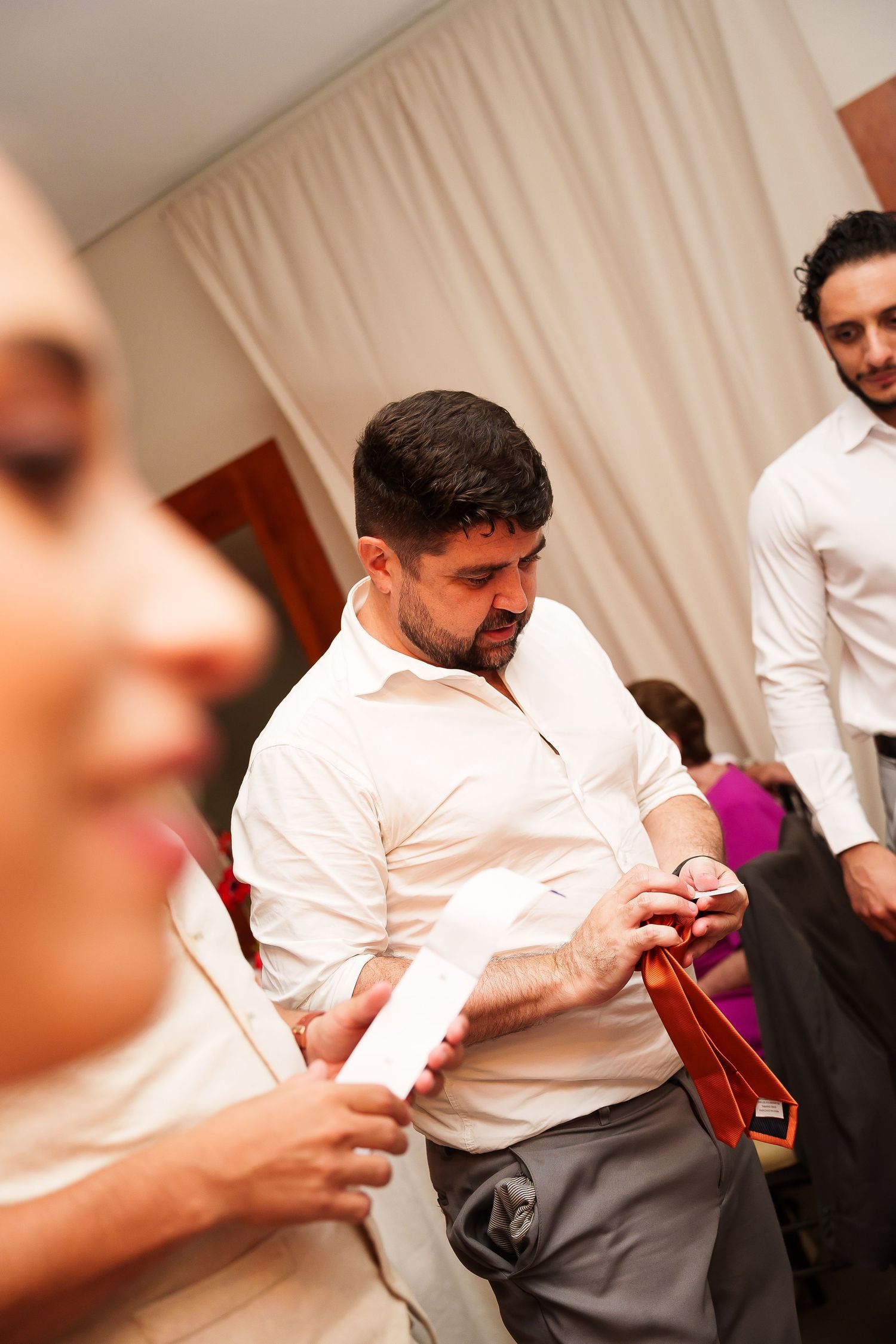 Man in white shirt holding a tie while getting dressed for a formal event indoors.